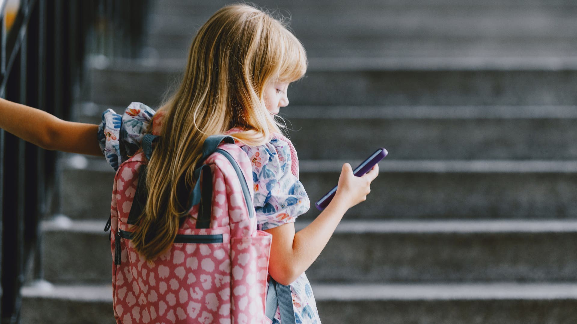 Ein Kind mit einem rosa Rucksack steht auf einer Treppe und schaut auf ein Smartphone. Es hält sich mit einer Hand am Geländer fest. Die Umgebung wirkt wie ein Schulgebäude.