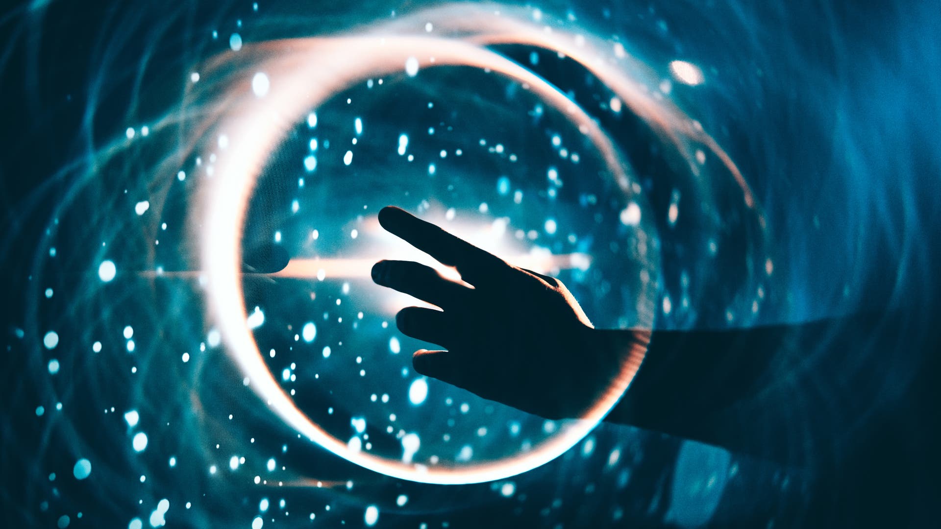 Eine Hand streckt sich in Richtung eines leuchtenden, kreisförmigen Lichts in einem dunklen, blauen Hintergrund. Um den Kreis herum sind helle, funkelnde Punkte verteilt, die an Sterne erinnern. Die Szene vermittelt ein Gefühl von Technologie und Magie.