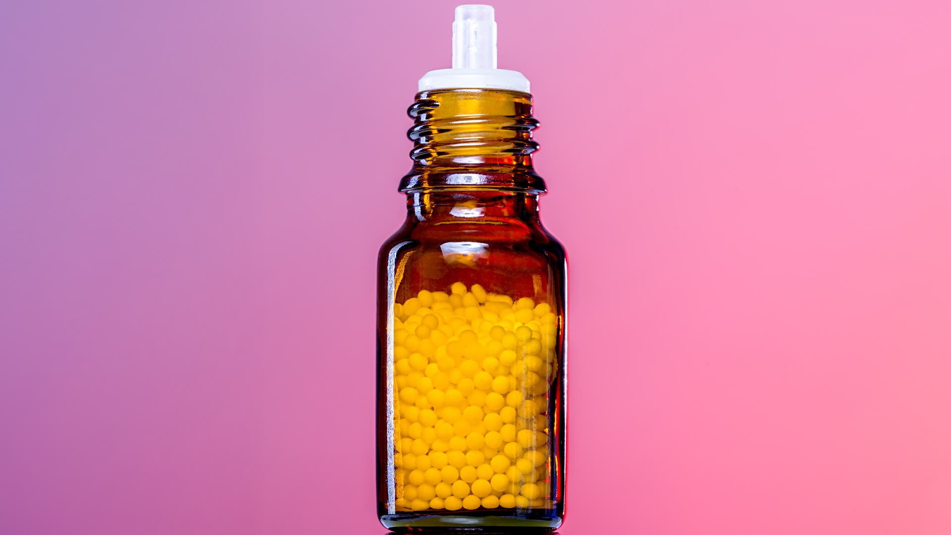 Eine braune Glasflasche mit einem weißen Verschluss, gefüllt mit homöopathischen Kügelchen. Der Hintergrund ist in einem sanften Rosa gehalten.