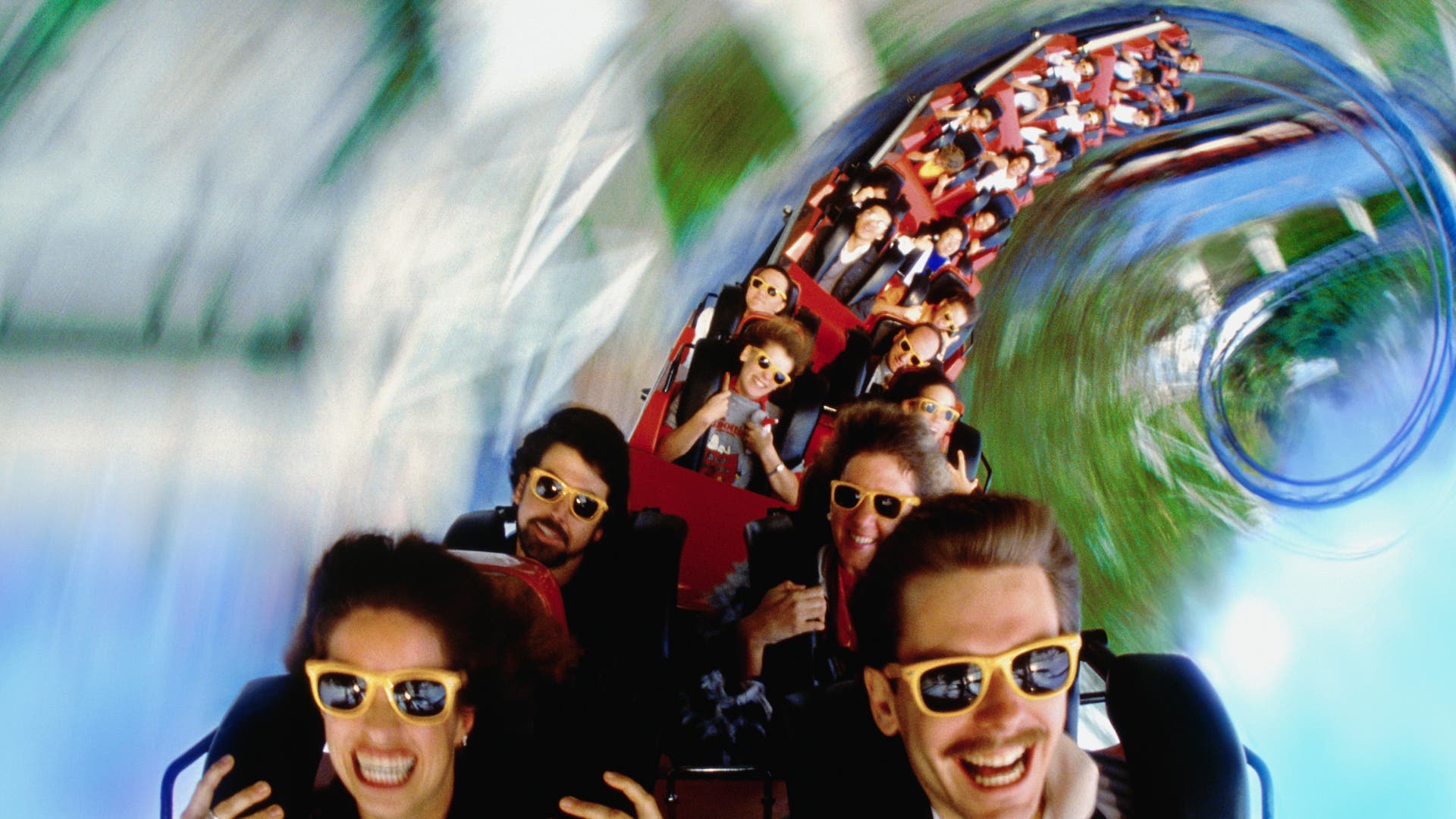 Menschen fahren in einem Achterbahnwagen durch eine Looping-Strecke. Sie tragen Sonnenbrillen und wirken begeistert und aufgeregt. Die Umgebung ist verschwommen, was die Geschwindigkeit und Dynamik der Fahrt betont. Die Szene vermittelt ein Gefühl von Abenteuer und Spaß.