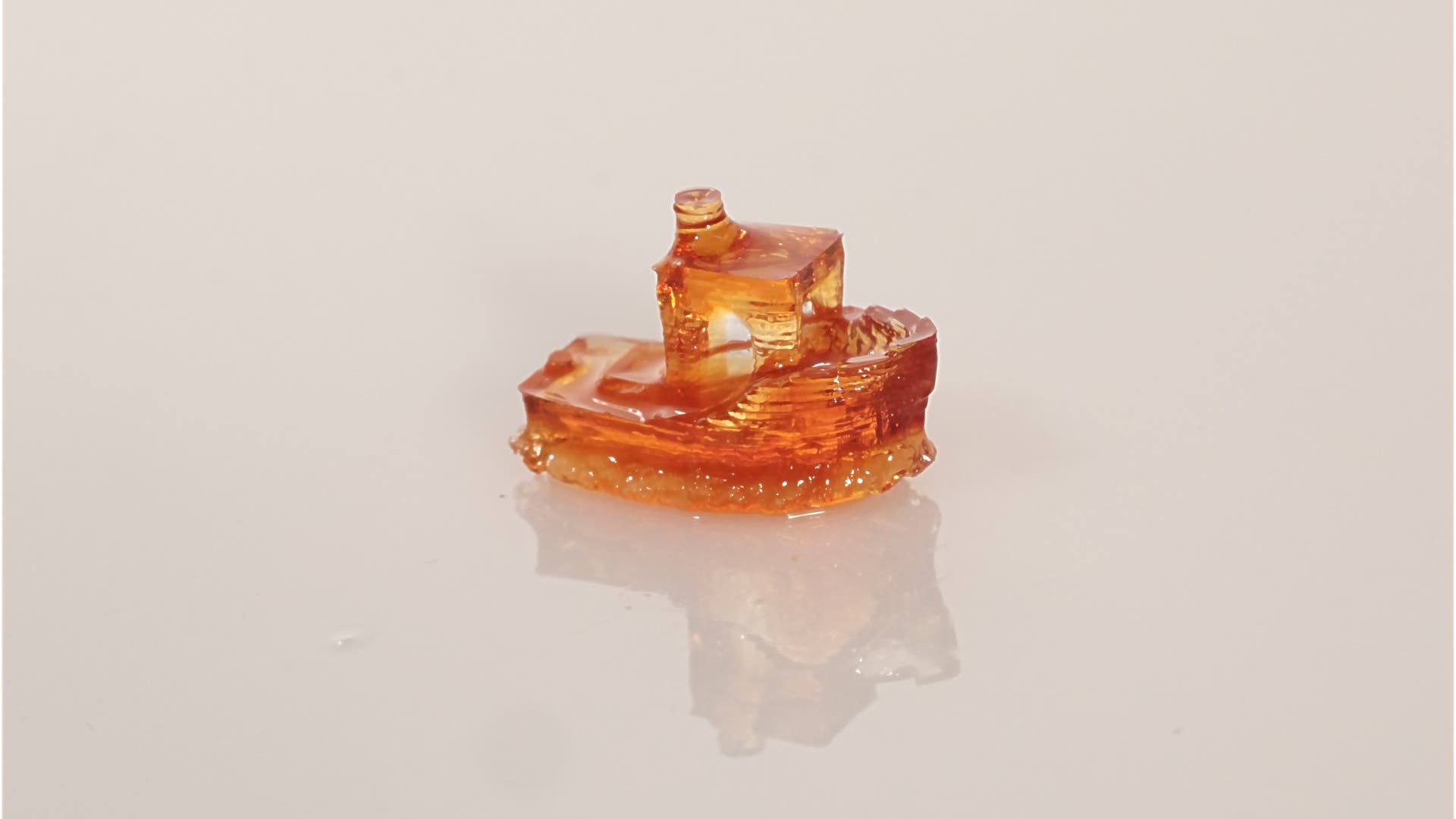 Ein kleines, transparentes, orangefarbenes 3-D-gedrucktes Modell eines Bootes auf einer glatten, reflektierenden Oberfläche. Das Modell zeigt feine Details wie ein Deck und einen Schornstein.