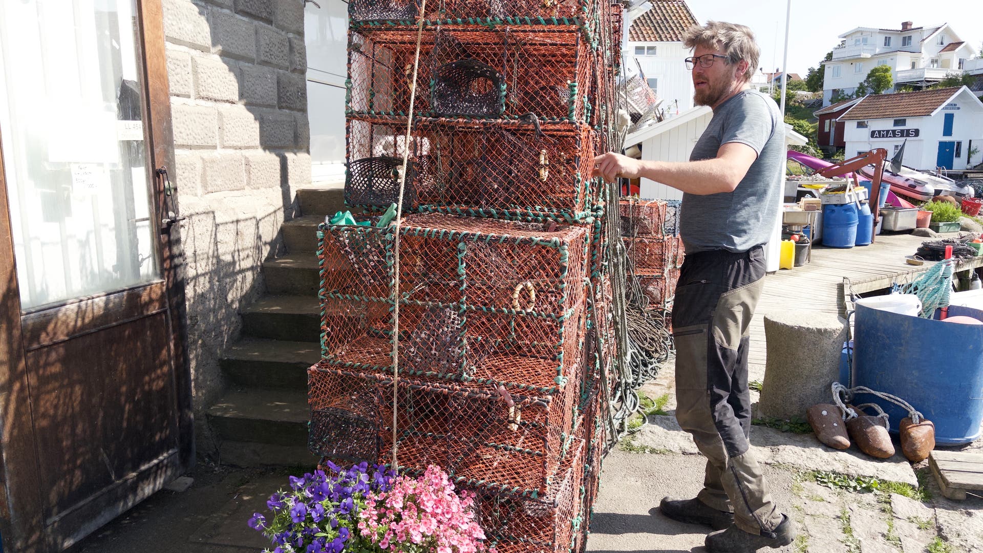 Ein Mann steht neben einem Stapel von Hummerkäfigen, die mit Seilen gesichert sind. Die Käfige sind aus Drahtgeflecht und befinden sich vor einem Gebäude mit Steinmauer. Im Vordergrund steht ein Blumentopf mit lila und rosa Blumen. Im Hintergrund sind weitere Gebäude und ein Hafenbereich zu sehen. Der Mann trägt ein graues T-Shirt und dunkle Hosen.