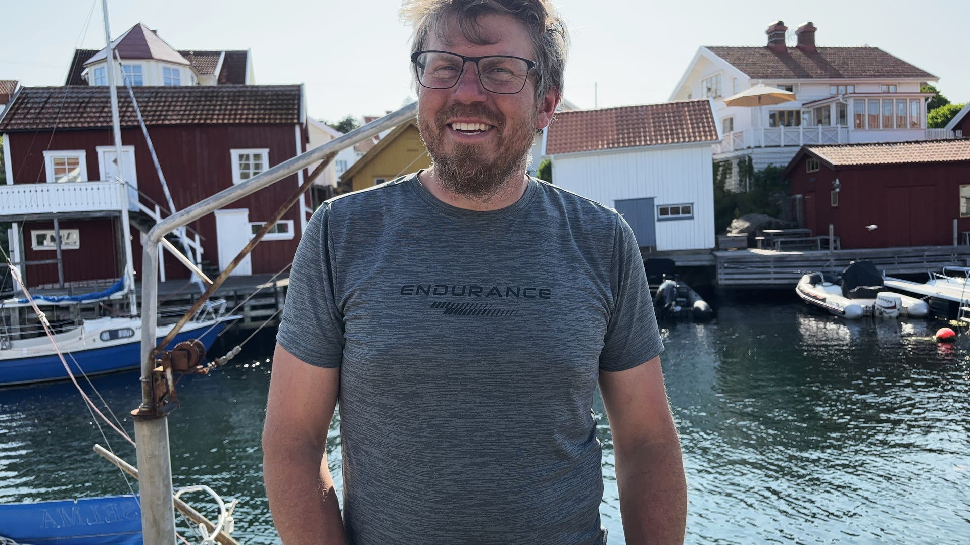 Ein Mann steht lächelnd vor einem Hafen mit mehreren Booten. Im Hintergrund sind bunte Holzhäuser zu sehen. Der Mann trägt ein graues T-Shirt mit der Aufschrift "ENDURANCE". Die Szene wirkt sonnig und friedlich.