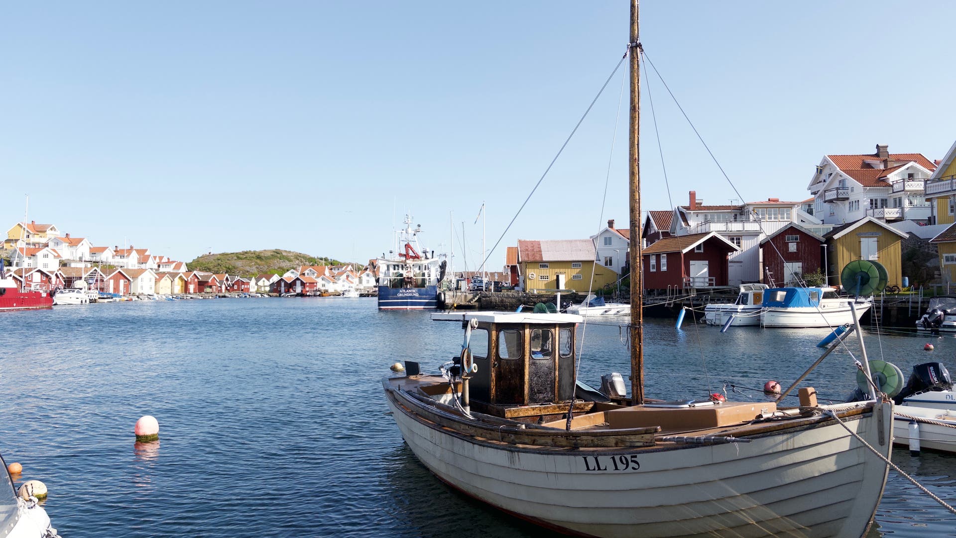 Ein traditionelles Holzboot mit der Kennzeichnung "LL 195" liegt in einem ruhigen Hafen vor Anker. Im Hintergrund sind bunte skandinavische Häuser und weitere Boote zu sehen. Der Himmel ist klar und blau, was auf einen sonnigen Tag hindeutet. Die Szene vermittelt eine friedliche und idyllische Atmosphäre in einer Küstenstadt.