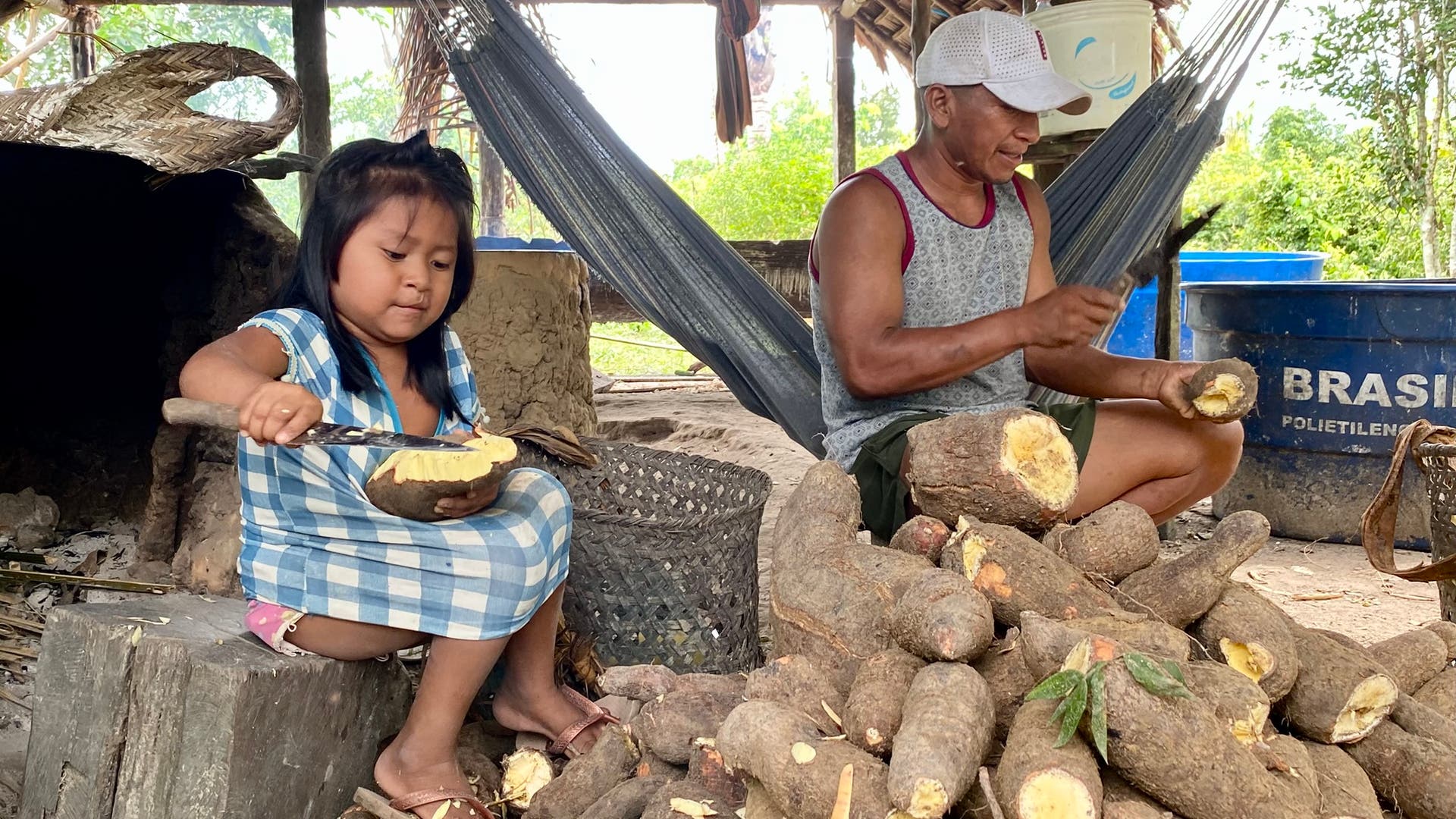 Ein Kind und ein Erwachsener sitzen in einer rustikalen Umgebung und schälen Maniokwurzeln. Das Kind trägt ein kariertes Kleid und sitzt auf einem Holzblock, während der Erwachsene auf einem Hocker sitzt und eine Mütze trägt. Beide sind von einem großen Haufen Maniokwurzeln umgeben. Im Hintergrund sind ein Hängemattenstuhl und ein blauer Behälter mit der Aufschrift "BRASIL" zu sehen. Die Szene vermittelt eine ländliche, handwerkliche Atmosphäre.