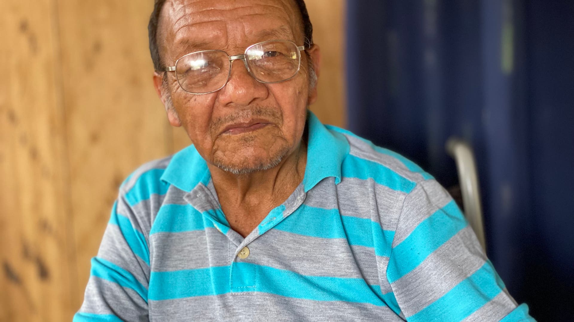Ein älterer Mann sitzt in einem Raum mit Holzwänden. Er trägt ein gestreiftes Poloshirt und schaut ruhig in die Kamera. Im Hintergrund ist ein blauer Vorhang zu sehen.