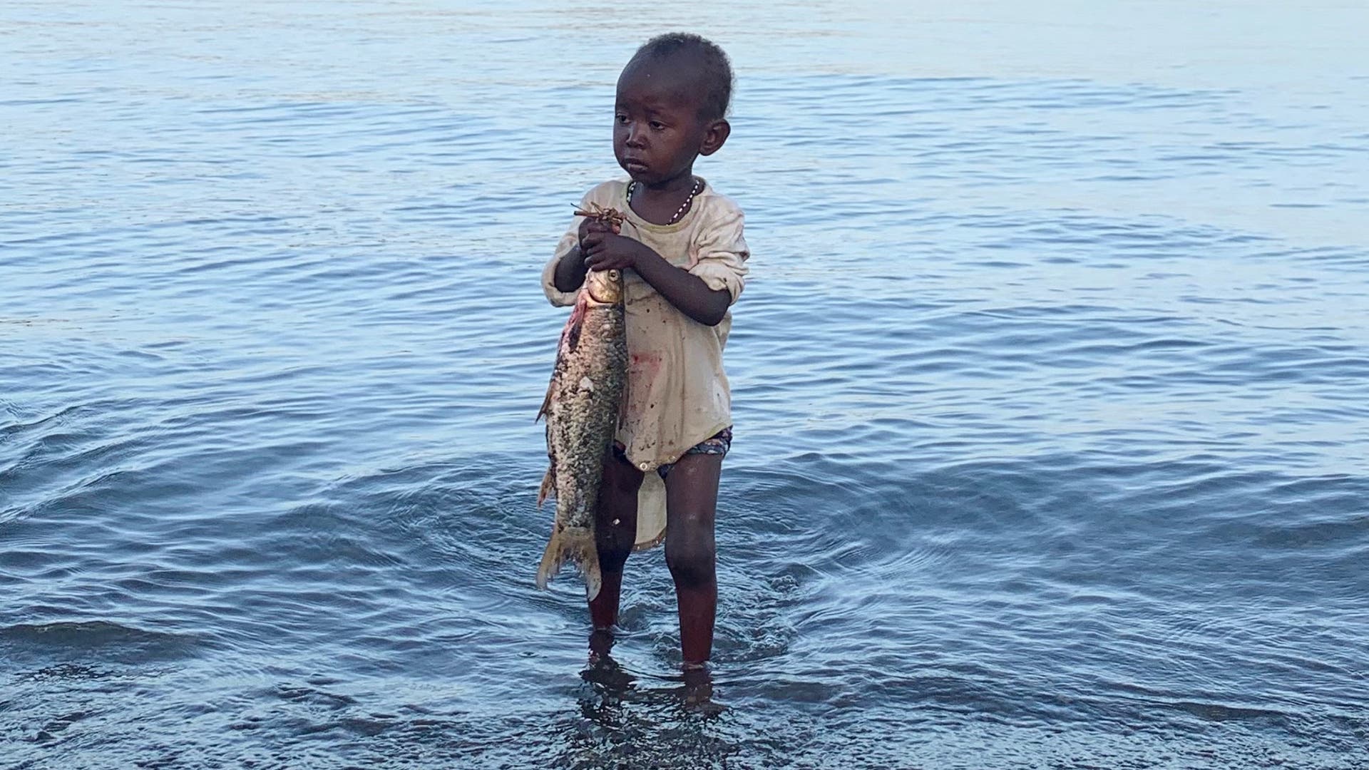 Ein Kleinkind steht im Wasser des Turkansees mit einem frisch gefangenen Fisch in der Hand.