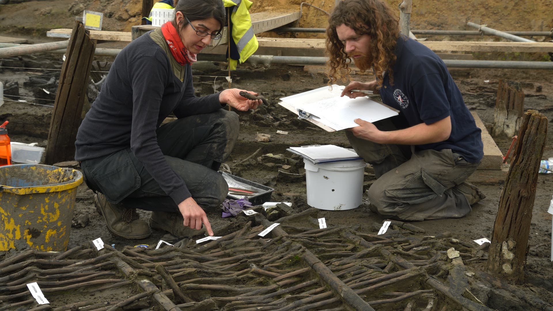 Zwei Archäologen arbeiten an einer Ausgrabungsstätte. Die Frau links untersucht ein Artefakt, während der Mann rechts Notizen auf einem Klemmbrett macht. Vor ihnen liegen freigelegte Holzstrukturen mit nummerierten Etiketten. Im Hintergrund sind Werkzeuge und Ausrüstungen sichtbar. Die Szene zeigt sorgfältige archäologische Arbeit und Dokumentation.