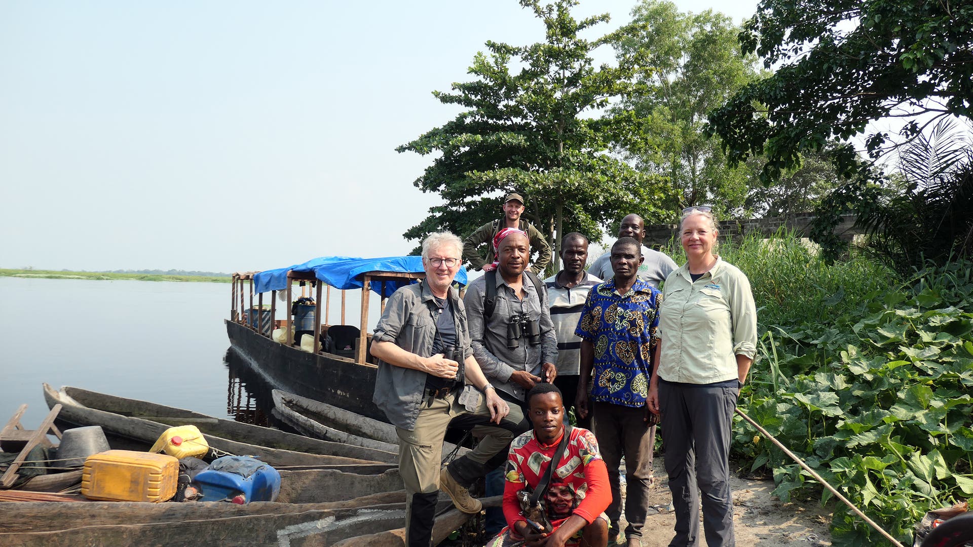 Eine Gruppe von Menschen steht am Ufer eines Flusses neben einem hölzernen Boot mit blauer Plane. Im Vordergrund sind traditionelle Kanus mit Kanistern und Taschen zu sehen. Die Personen lächeln in die Kamera, umgeben von üppigem Grün und Bäumen. Der Himmel ist klar und sonnig.