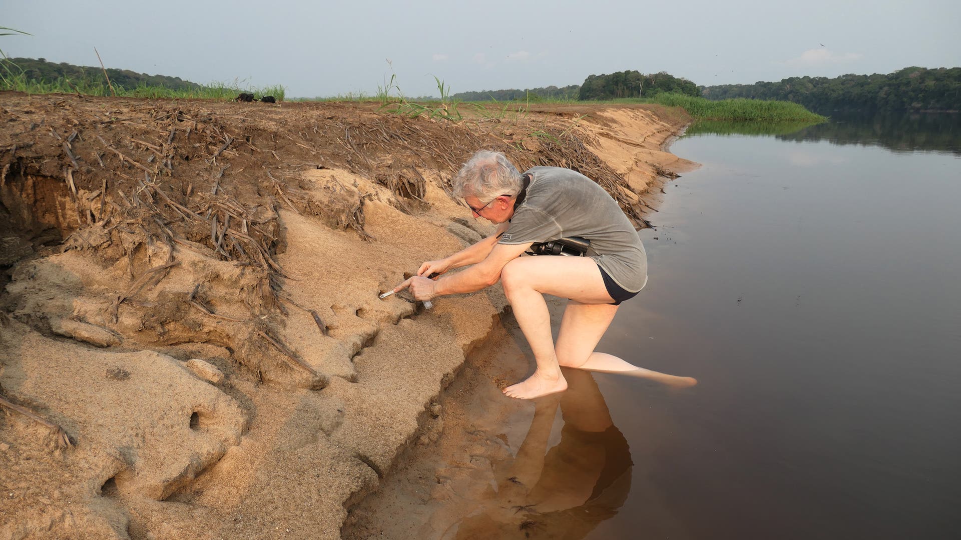 Eine Person mit grauem Haar kniet am Ufer eines Flusses und untersucht den sandigen Boden mit einem kleinen Werkzeug. Der Fluss spiegelt den klaren Himmel wider, und im Hintergrund sind Bäume und Vegetation zu sehen. Die Person trägt ein graues T-Shirt und Shorts und ist barfuß. Die Szene vermittelt eine ruhige, naturverbundene Atmosphäre.