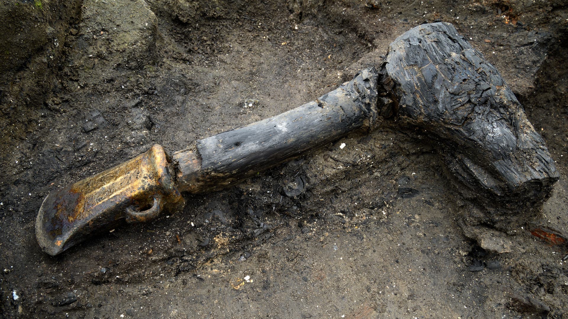 Ein gut erhaltenes, spätbronzezeitliches Beil, das in einem archäologischen Ausgrabungsgebiet im Boden liegt. Der Holzgriff ist dunkel und zeigt deutliche Spuren von Alterung und Verwitterung. Der Kopf des Werkzeugs ist aus Bronze und weist eine abgenutzte Oberfläche auf. Die Umgebung besteht aus erdigem Boden mit kleinen Steinen und Schmutzpartikeln.