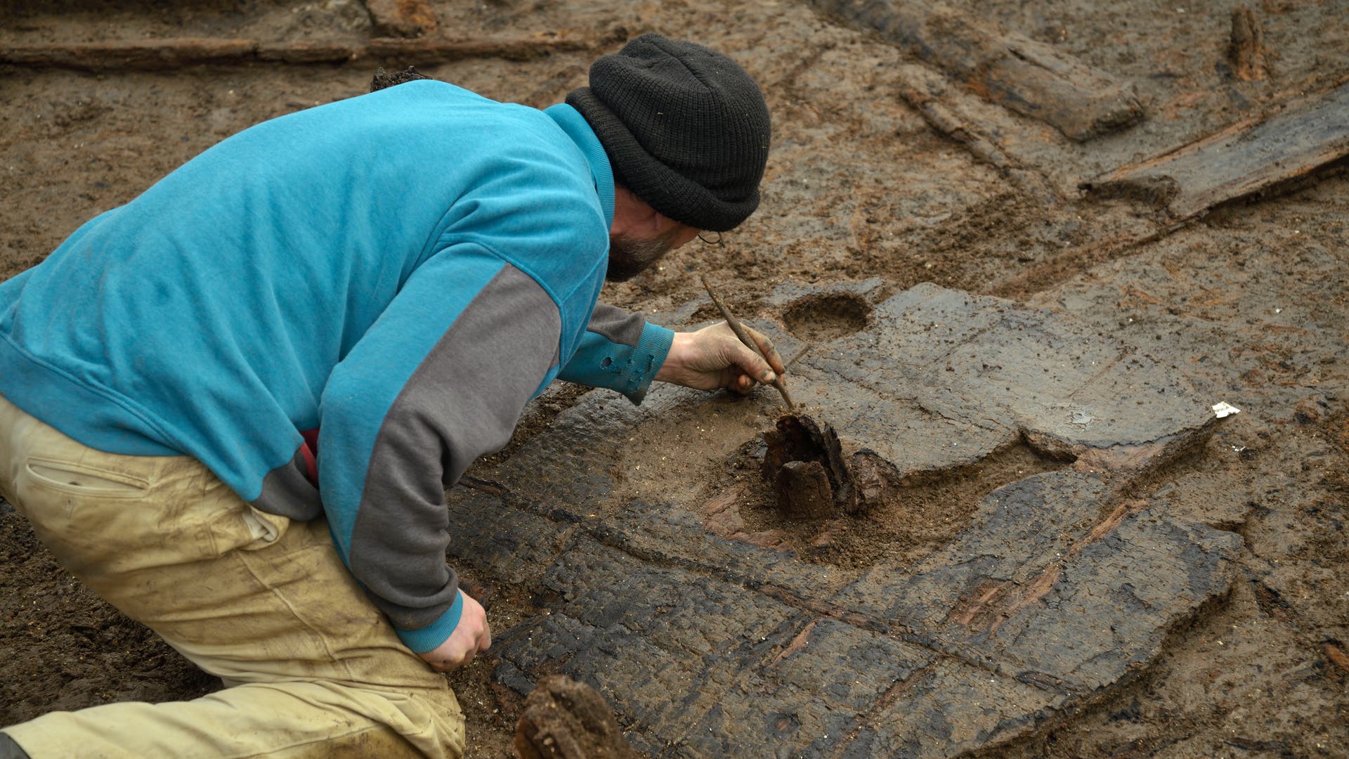 Ein Archäologe in blauer Jacke und schwarzer Mütze kniet auf einem Ausgrabungsfeld und verwendet ein kleines Werkzeug, um vorsichtig Erde von einem großen, flachen Artefakt zu entfernen. Die Umgebung ist schlammig und es sind Holzstrukturen im Boden sichtbar.