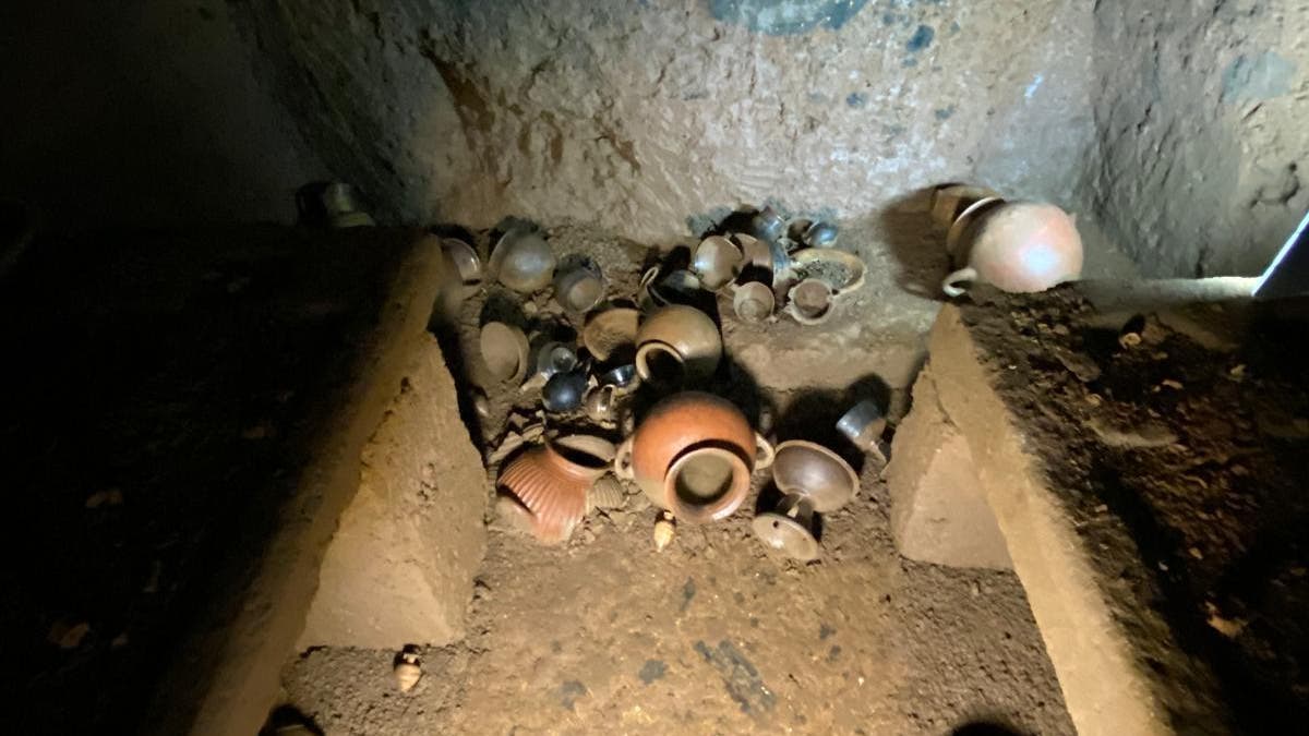 Ein unterirdischer Raum mit mehreren antiken Tongefäßen, die auf dem Boden verstreut liegen. Die Wände sind aus grobem Stein mit blauen Verfärbungen. Das Bild zeigt eine archäologische Ausgrabungsstätte, die von einer Lichtquelle beleuchtet wird, um die Details der Keramik und der Umgebung hervorzuheben.