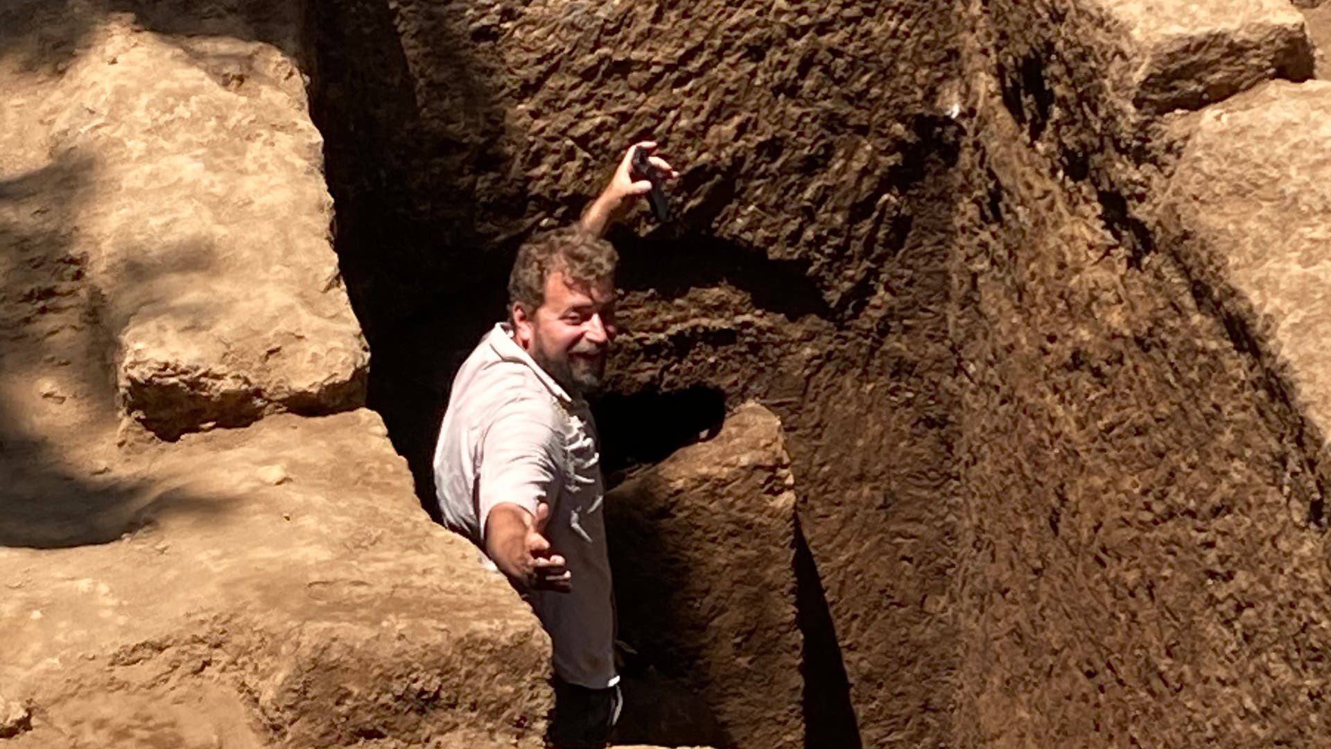 Eine Person steht in einer tiefen archäologischen Ausgrabungsstätte, umgeben von Erdschichten. Sie hält ein Werkzeug in der Hand und blickt nach oben. Eine Leiter ist am Rand der Grube sichtbar. Die Szene ist von Sonnenlicht beleuchtet, das die Textur der Erde hervorhebt.