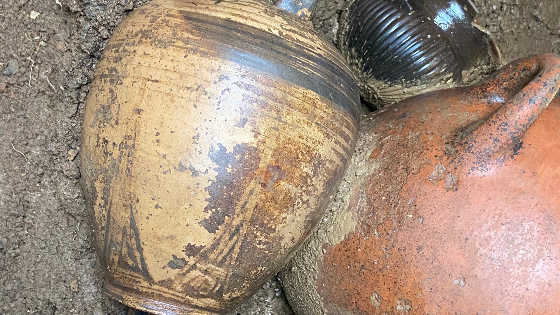 Eine archäologische Ausgrabung zeigt mehrere antike Keramikgefäße, die teilweise im Erdreich eingebettet sind. Ein großes, verziertes Gefäß liegt im Vordergrund, während kleinere, dunklere Gefäße im Hintergrund sichtbar sind. Die Gefäße weisen unterschiedliche Formen und Oberflächenmuster auf, die auf ihre historische Nutzung hinweisen könnten. Der Boden ist feucht und erdig, was auf die Ausgrabungsbedingungen hindeutet.