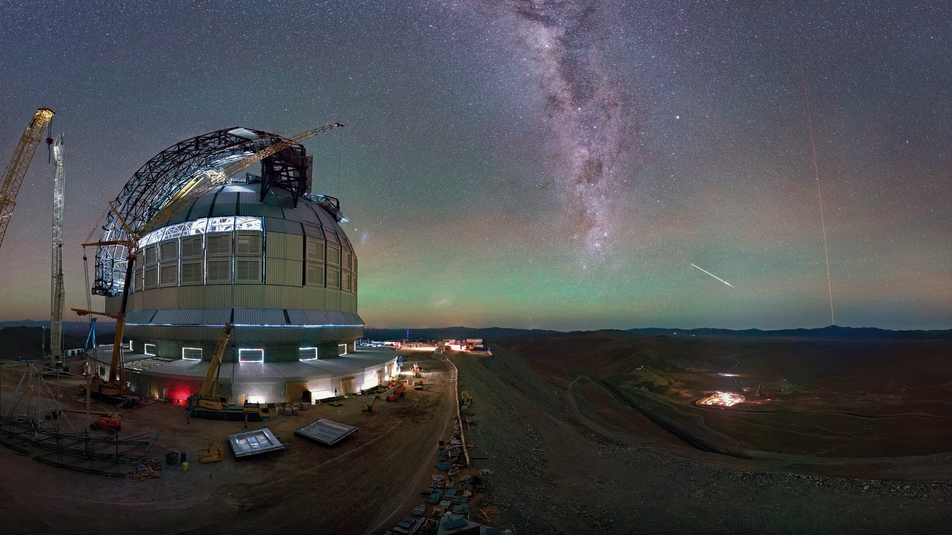 Ein großes Observatorium mit einer Kuppel und Baukränen steht in einer Wüstenlandschaft unter einem klaren Nachthimmel. Die Milchstraße ist deutlich sichtbar, und ein heller Meteor zieht eine Linie am Himmel. Im Hintergrund sind Berge und Lichter einer entfernten Siedlung zu sehen. Die Szene vermittelt eine ruhige, aber beeindruckende Atmosphäre der Astronomie und Technik.