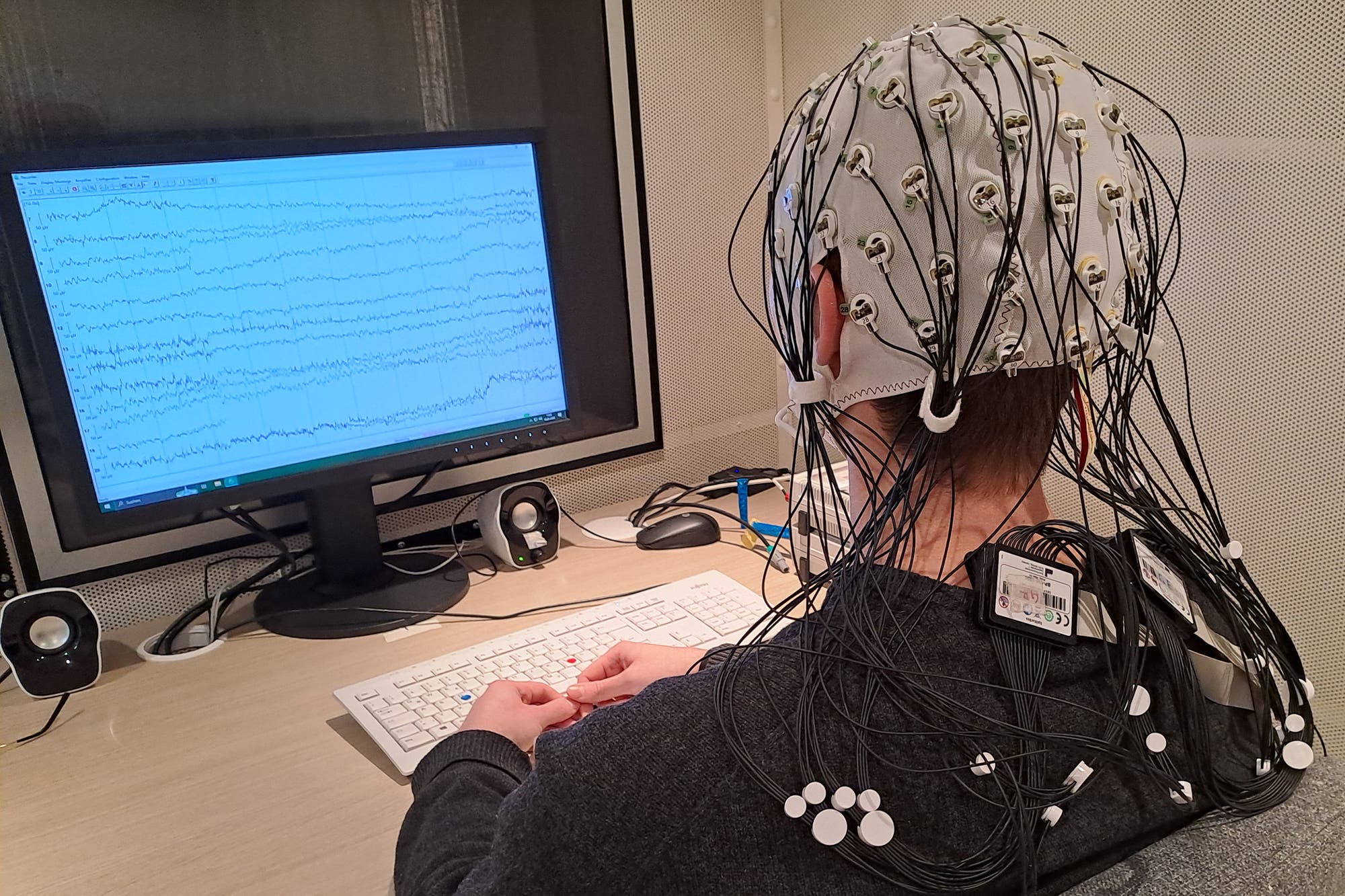 Eine Person sitzt an einem Schreibtisch vor einem Computerbildschirm, der EEG-Daten mit Wellenmustern anzeigt. Sie trägt eine EEG-Haube mit zahlreichen Elektroden und Kabeln, die an der Haube befestigt sind; ihre Hände ruhen auf einer Computertastatur. Der Raum ist mit schallabsorbierenden Paneelen ausgestattet, und neben dem Monitor stehen zwei Lautsprecher.