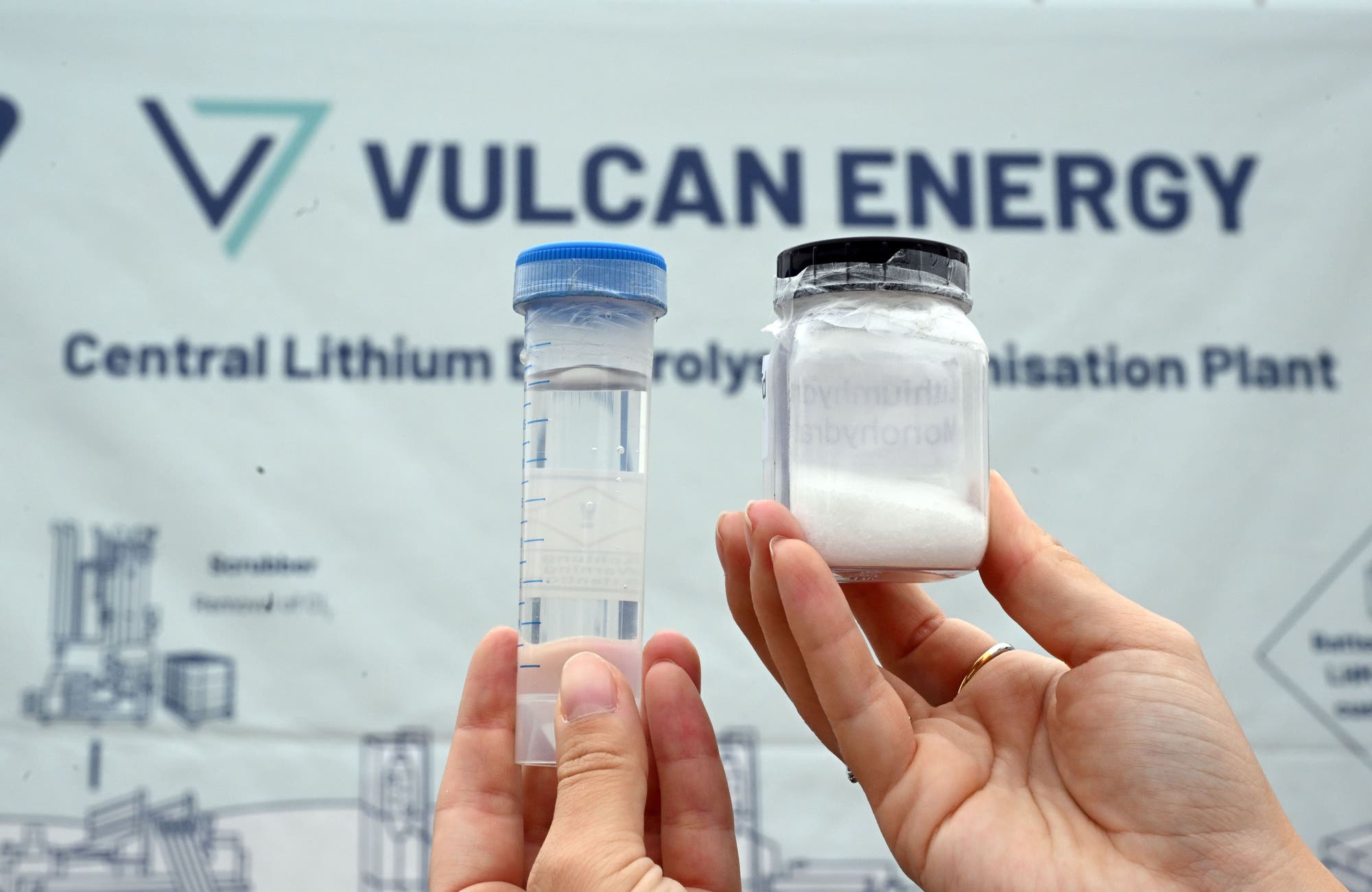 Zwei Hände halten ein Reagenzglas mit klarer Flüssigkeit und ein Glas mit weißem Pulver vor einem Schild mit der Aufschrift "Vulcan Energy Central Lithium Electrolysis Decarbonisation Plant". Im Hintergrund sind schematische Darstellungen von Anlagen zu sehen. Das Bild zeigt Materialien, die möglicherweise in einem industriellen oder wissenschaftlichen Kontext verwendet werden.