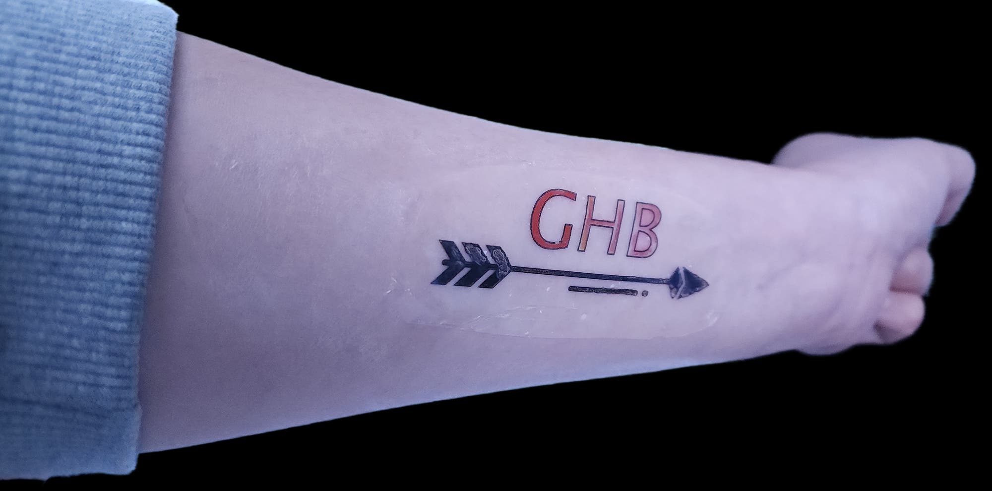 Ein Unterarm mit einem Tattoo, das einen schwarzen Pfeil und die Buchstaben "GHB" in Rot zeigt. Der Pfeil ist vertikal ausgerichtet, mit der Spitze nach oben. Der Arm ist in einem neutralen Hintergrund positioniert.