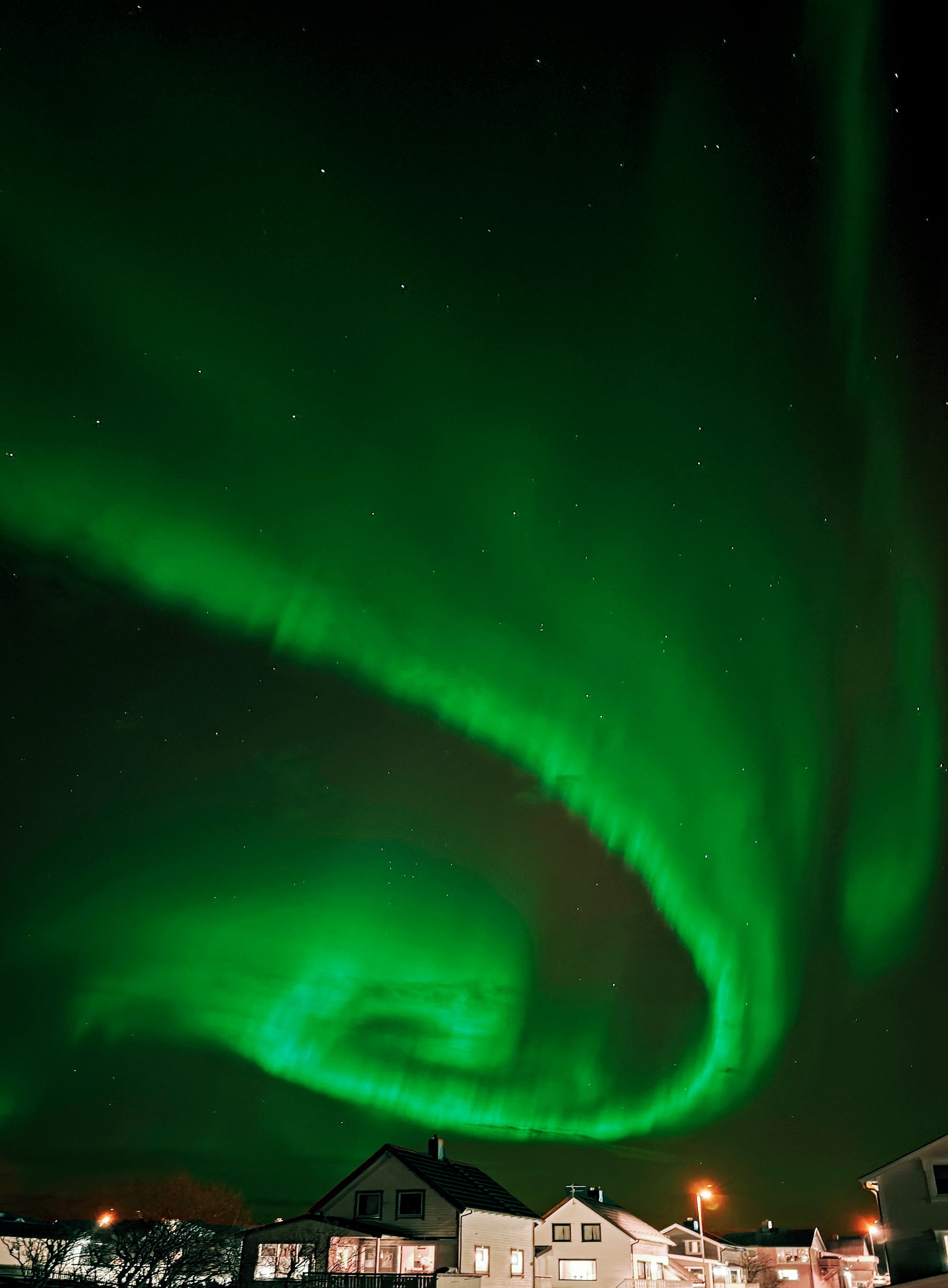 Ein nächtlicher Himmel mit leuchtendem grünem Polarlicht, das sich in geschwungenen Mustern über den Sternen erstreckt. Unten sind die Umrisse von Häusern zu sehen, die im Dunkeln stehen. Das Bild zeigt die Aurora Borealis, ein beeindruckendes Naturphänomen, das durch die Interaktion von Sonnenwinden mit der Erdatmosphäre entsteht.