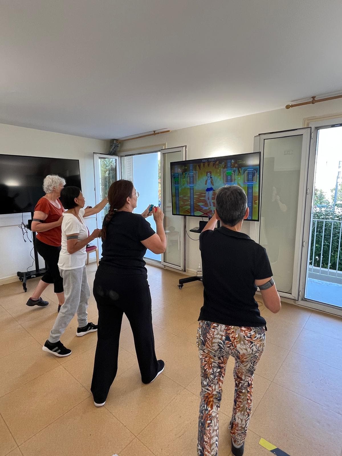 Vier Frauen stehen in einem hellen Raum und reagieren auf die Bewegungen der Figur auf dem Bildschirm. Der Raum hat große Fenster, die viel Tageslicht hereinlassen. Eine Person trägt ein rotes Oberteil, die anderen sind in dunkleren Farben gekleidet. 