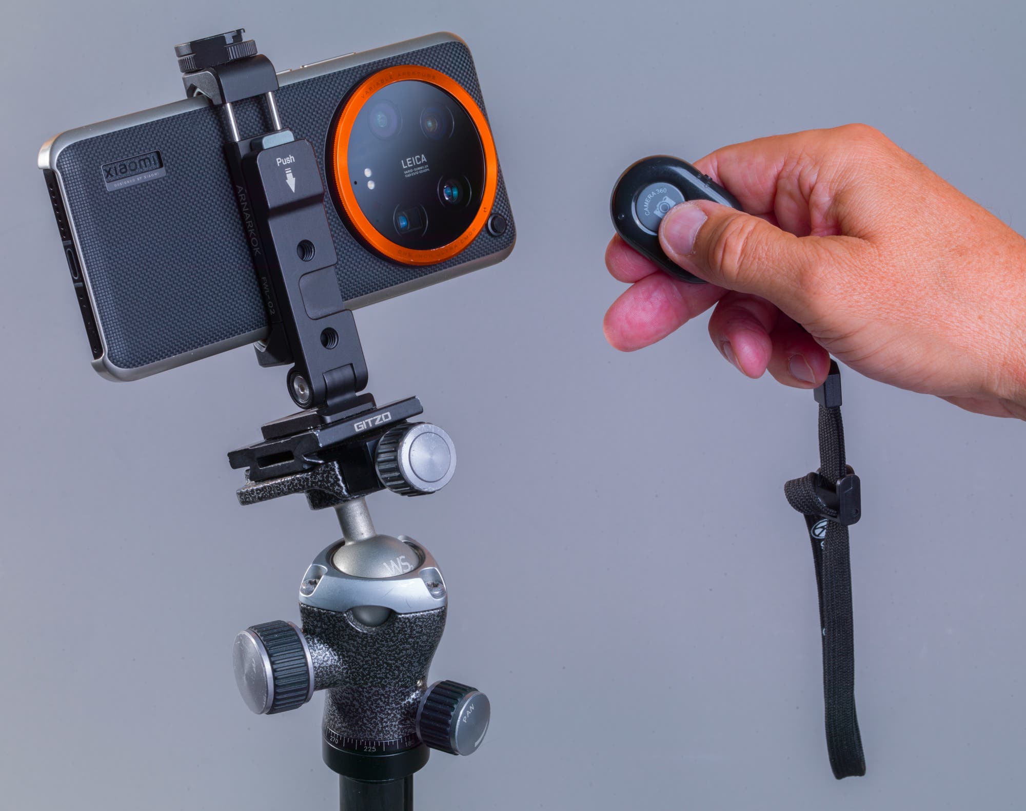 Ein Smartphone ist auf einem Stativ montiert. Eine Hand hält eine Fernbedienung, um das Gerät auszulösen. Das Smartphone hat eine auffällige Kamera mit einem orangefarbenen Ring. Der Hintergrund ist neutral grau.