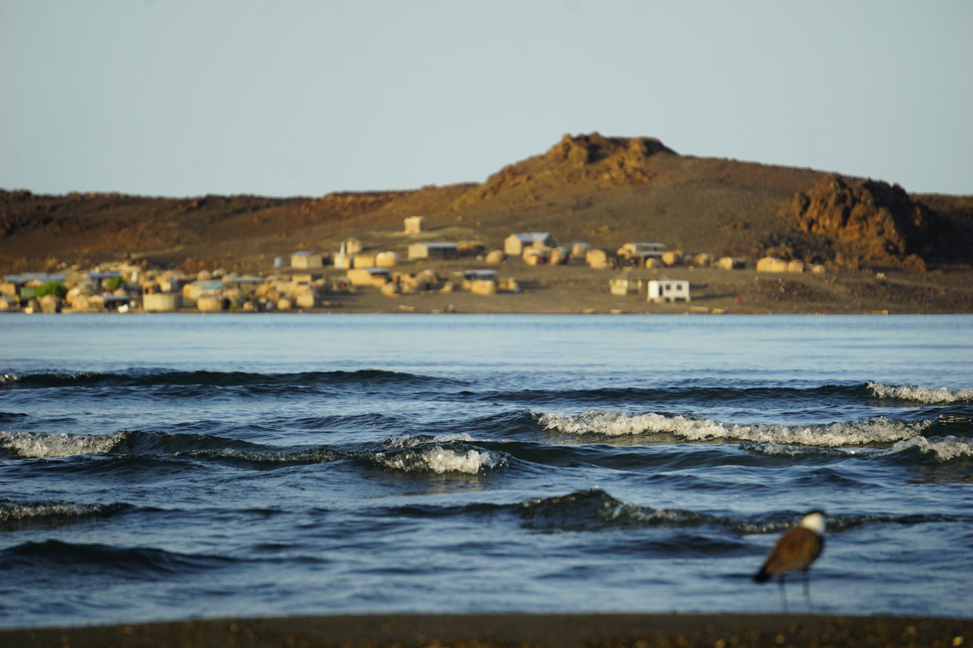 Der Turkanasee schlägt leichte Wellen im Vordergrund des Bilds. Im Hintergrund sind Hütten der El Molo unscharf zu erkennen.