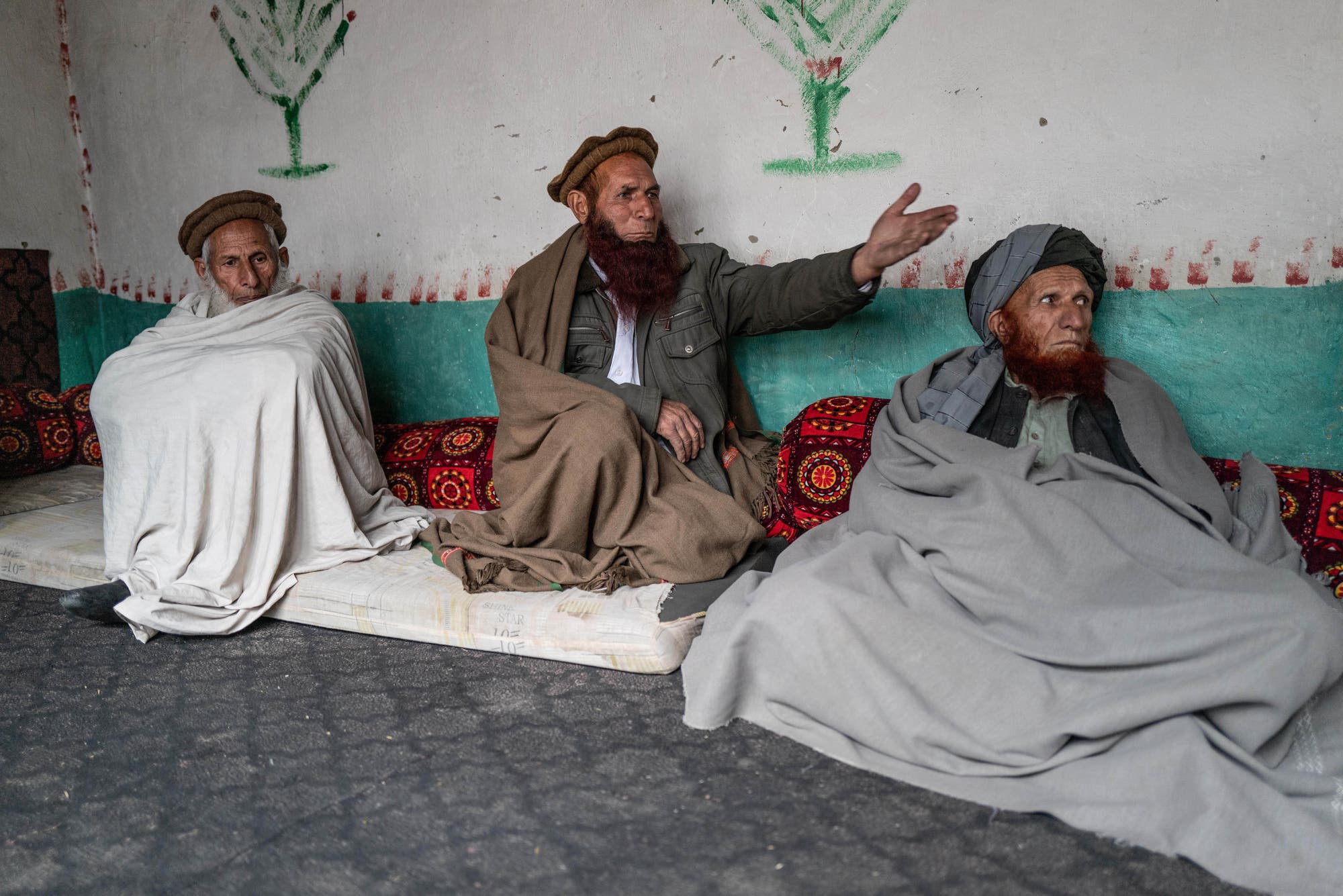 Drei Männer sitzen auf Matratzen auf dem Biden und sind in Decken eingehüllt. 