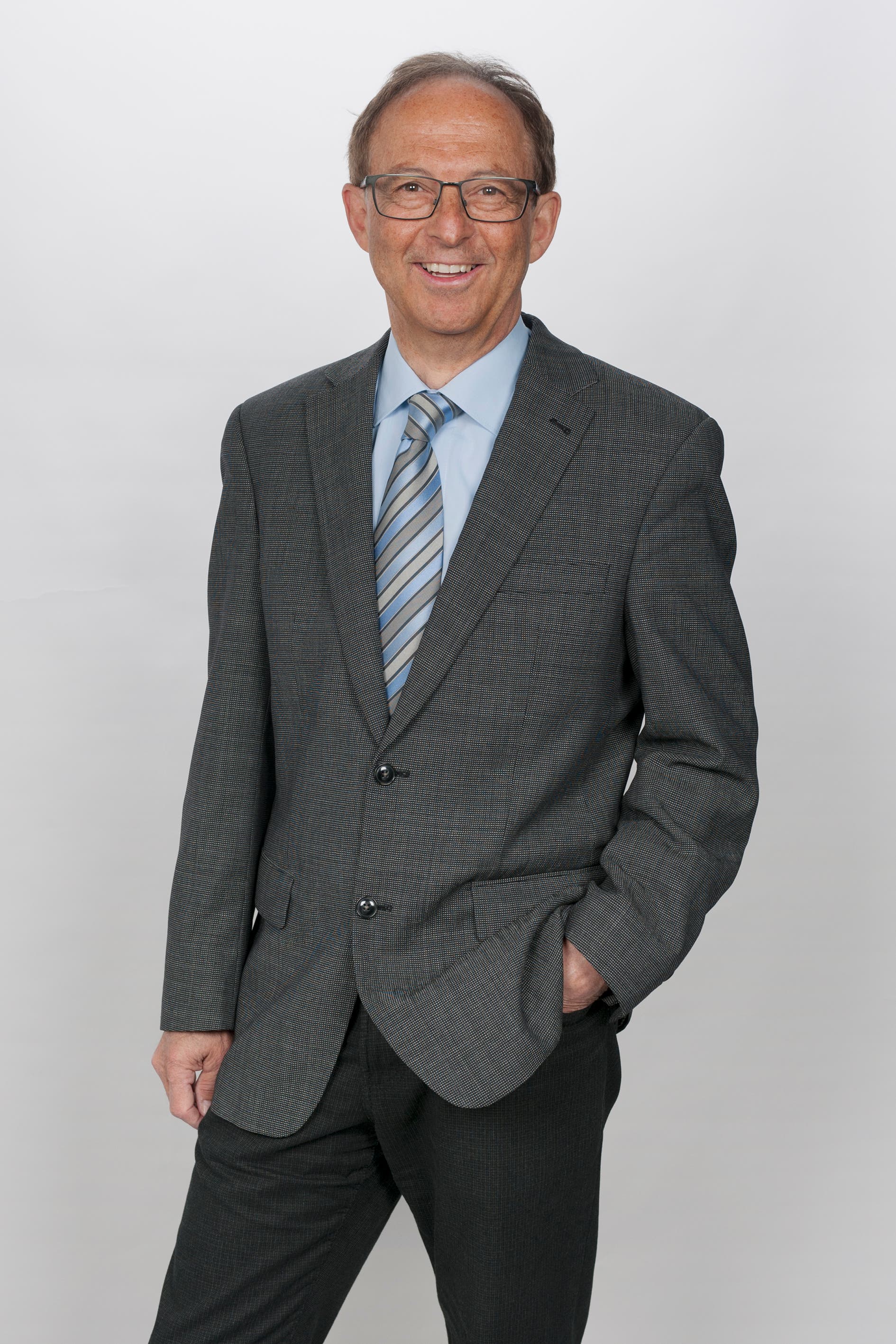 Ein Mann in einem grauen Anzug mit Krawatte steht vor einem neutralen Hintergrund. Er lächelt in die Kamera und hat eine Hand in der Hosentasche.