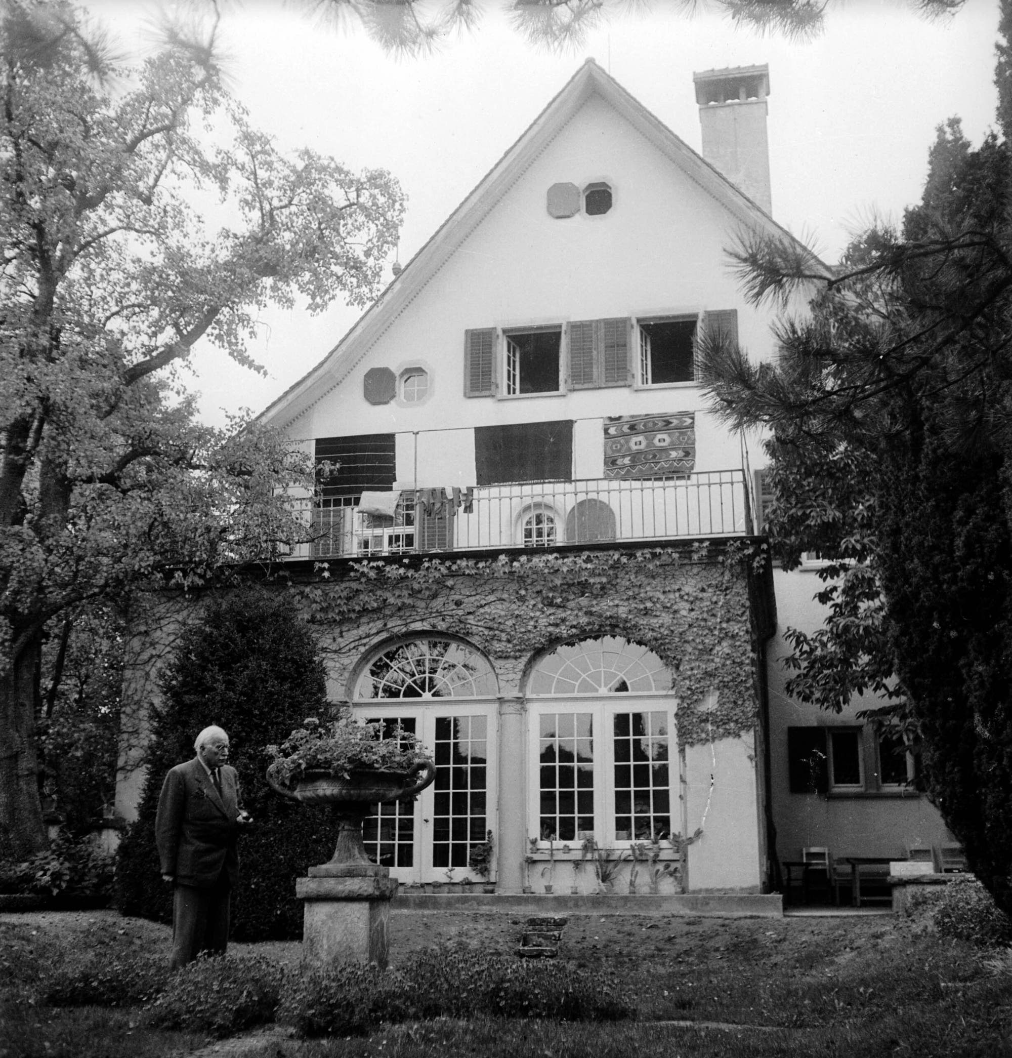 Ein Mann mit weißem Haar steht in einem gepflegten Garten vor einem großen, zweistöckigen Haus mit Satteldach. Die Fassade des vorgelagerten Wintergartens ist mit Efeu bewachsen, und auf dem Balkon darüber hängen Teppiche oder Decken. Der Mann betrachtet einen dekorativen Steinvogelbrunnen. Im Hintergrund sind hohe Bäume zu sehen, die das Haus umgeben.