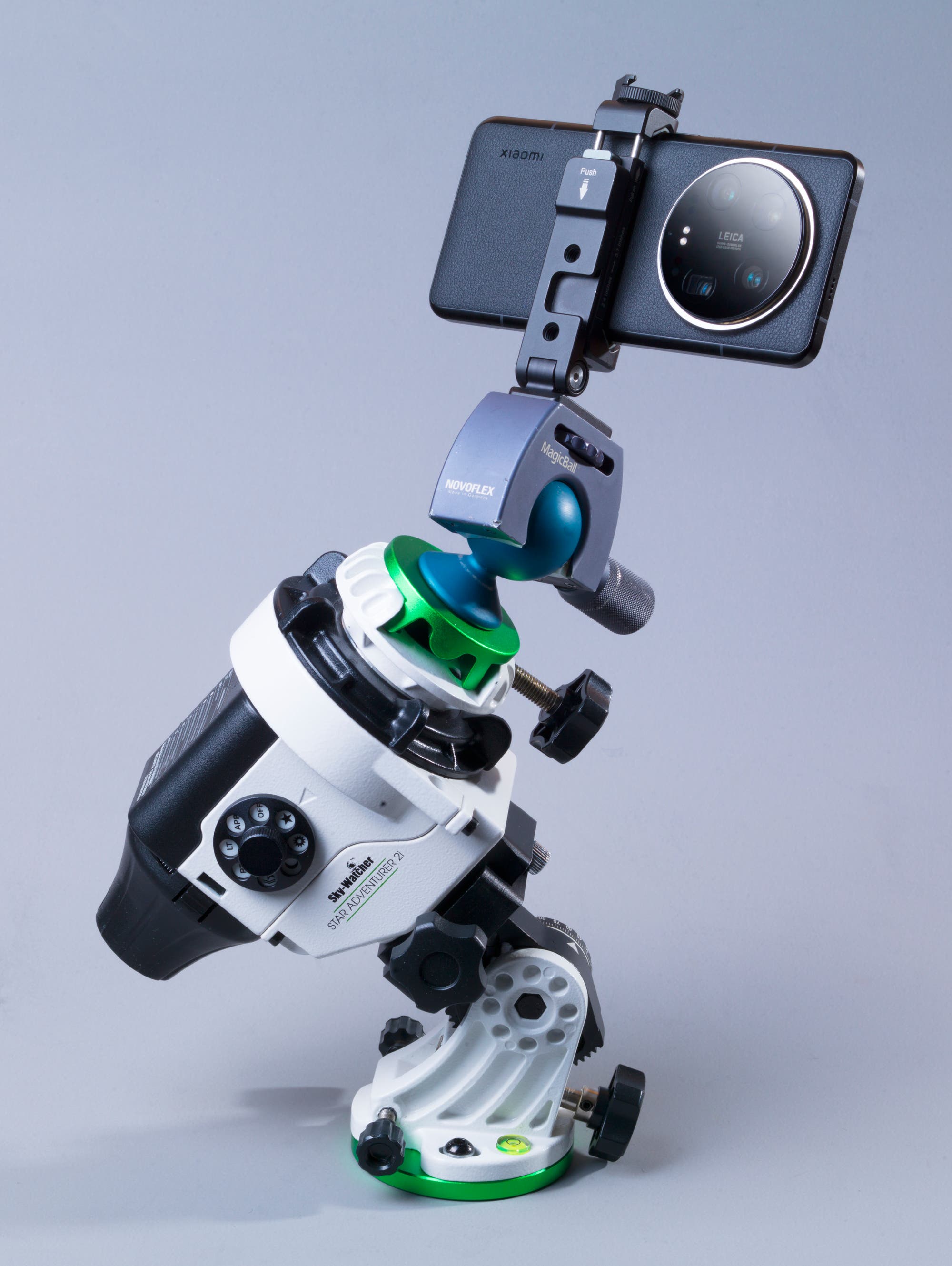 Ein Smartphone ist auf einem Teleskop-Montierungssystem befestigt. Das Teleskop ist schräg positioniert und zeigt verschiedene Einstellknöpfe und -hebel. Das Smartphone ist mit einer Halterung verbunden, die es in einer stabilen Position hält. Auf dem Smartphone sind die Marken "Xiaomi" und "Leica" sichtbar. Die Szene zeigt eine Kombination aus moderner Technologie und optischen Geräten, die für Fotografie oder Beobachtungen verwendet werden könnte.