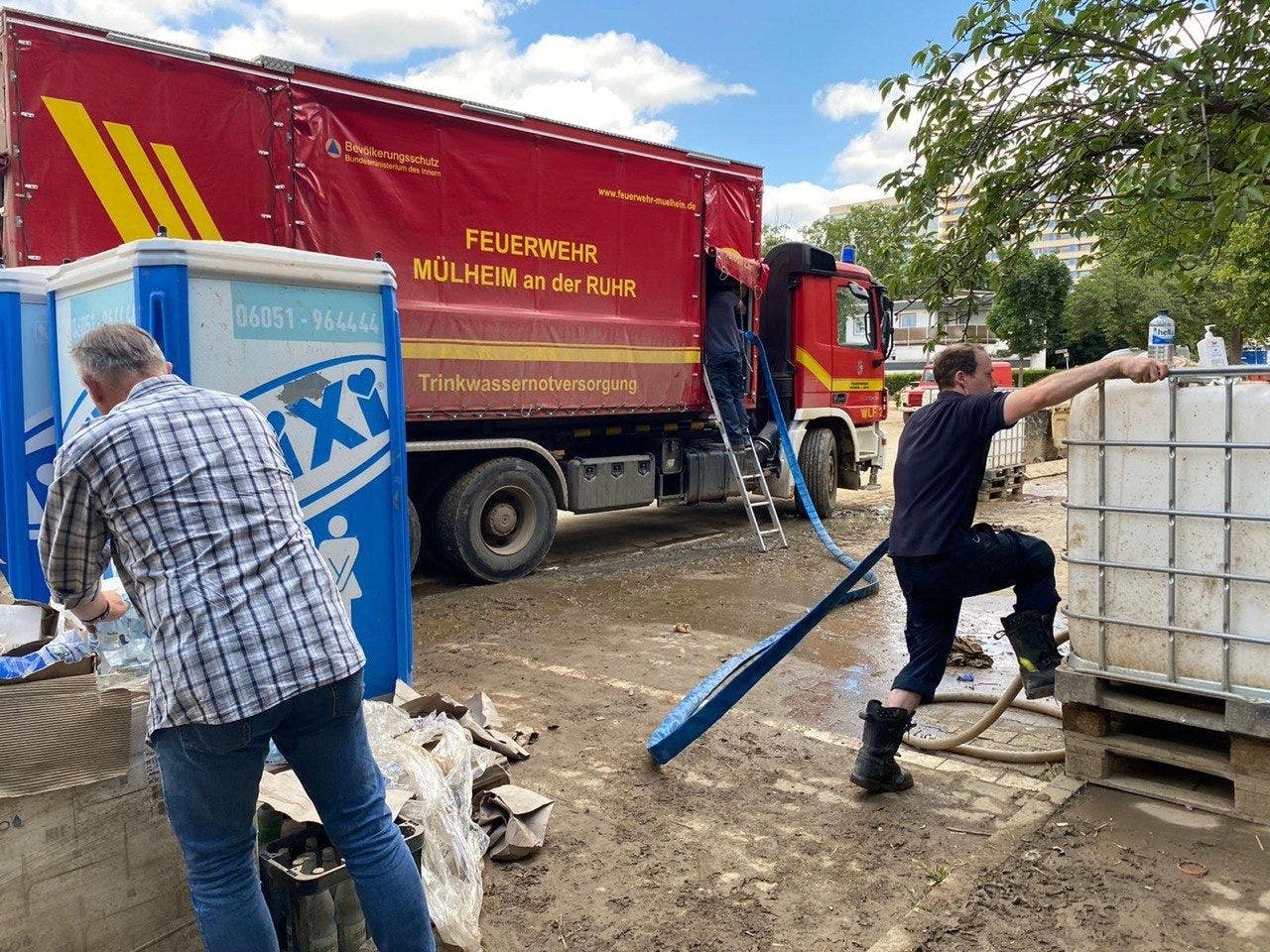 Ein Feuerwehrfahrzeug der Feuerwehr Mülheim an der Ruhr steht auf einer schlammigen Straße. Zwei Feuerwehrleute sind beschäftigt: Einer befestigt einen blauen Schlauch an einem großen Wassertank, während der andere auf eine Leiter steigt, die zum Lkw führt. Ein Mann im karierten Hemd steht links und arbeitet an einem blauen mobilen Toilettenhäuschen. Der Himmel ist teilweise bewölkt. Text auf dem Lkw: "FEUERWEHR MÜLHEIM AN DER RUHR" und "Trinkwassernotversorgung".
