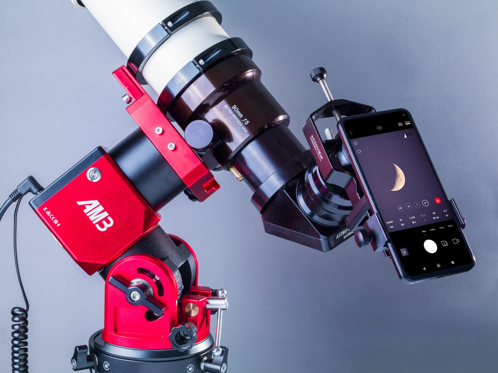 Ein Teleskop mit einer roten Montierung ist mit einem Smartphone verbunden, das ein Bild des Mondes auf dem Bildschirm zeigt. Das Teleskop hat eine 80-mm-<em>f</em>6-Linse. Das Smartphone ist in einer Halterung befestigt und zeigt eine App zur Astrofotografie. Die Szene illustriert die Verbindung von moderner Technologie mit traditioneller Astronomie.