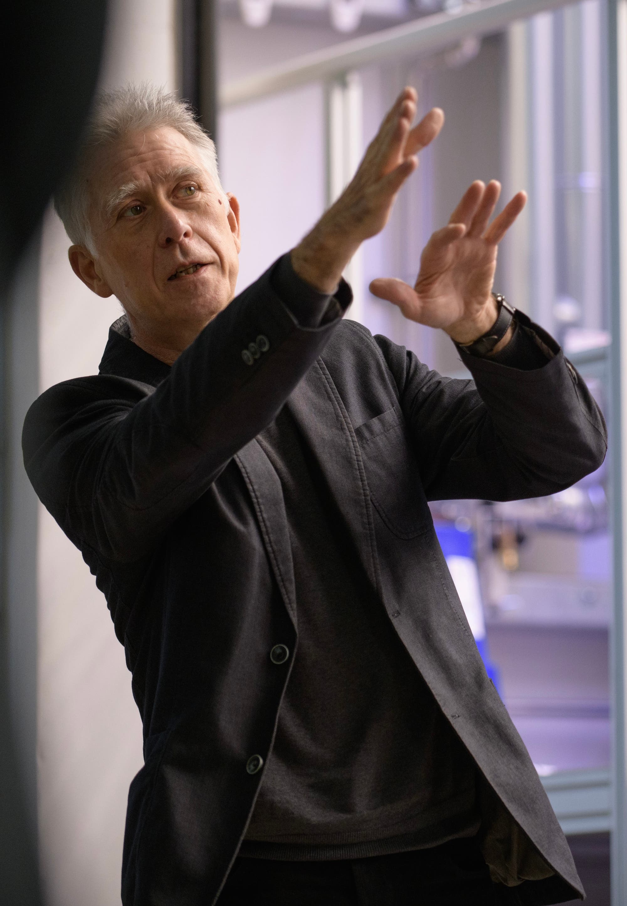 Eine Person in einem dunklen Anzug gestikuliert mit den Händen, als ob sie etwas erklärt oder diskutiert. Der Hintergrund zeigt unscharfe technische Geräte, was auf eine wissenschaftliche oder technische Umgebung hindeutet. Die Person scheint in ein Gespräch vertieft zu sein.