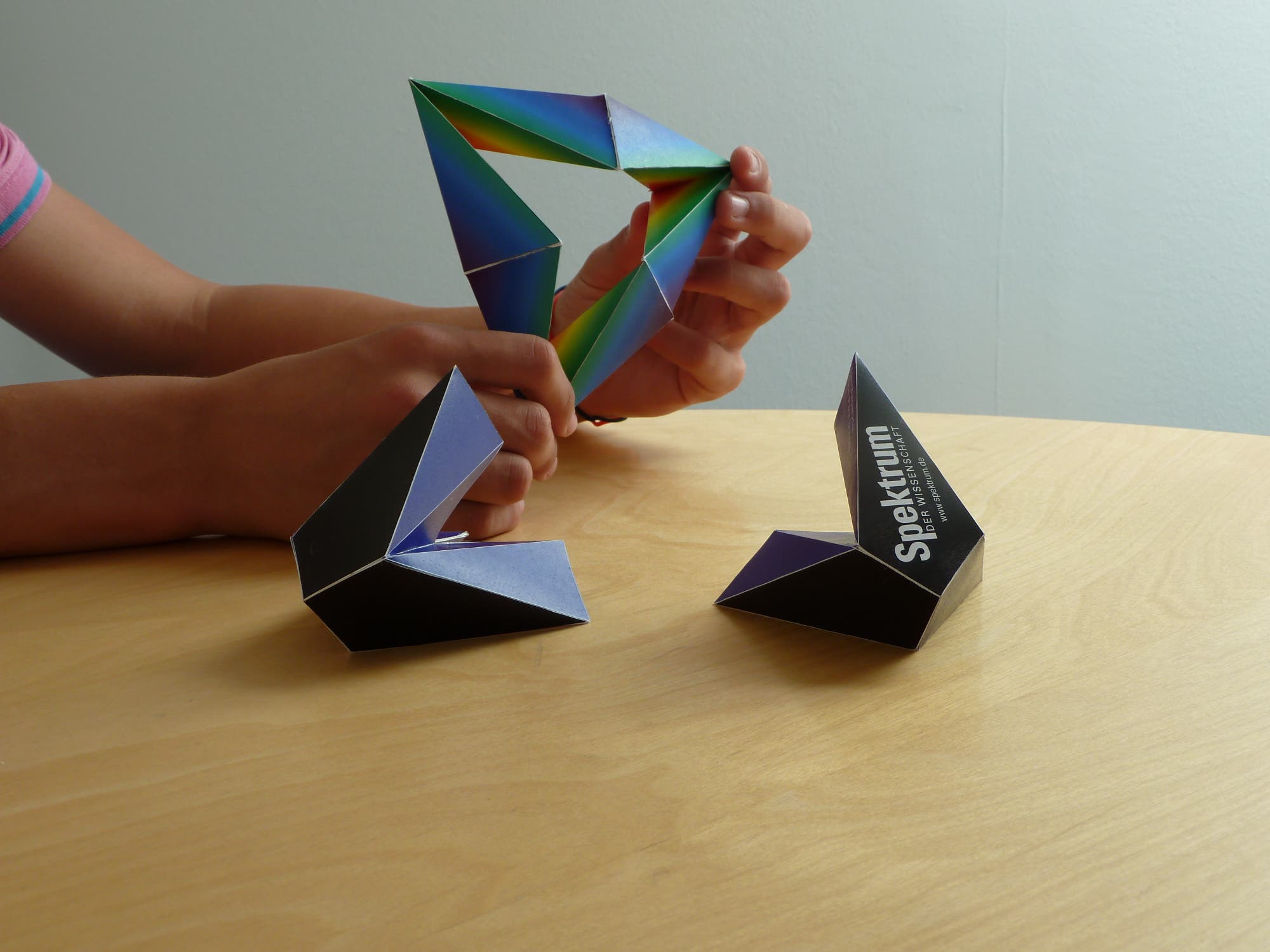 Eine Person formt ein buntes, dreieckiges Origami auf einem Holztisch. Zwei weitere Origami-Strukturen sind auf dem Tisch platziert. Auf einem der Origamis steht der Text "Spektrum". Die Szene zeigt kreative Handarbeit mit Papier.