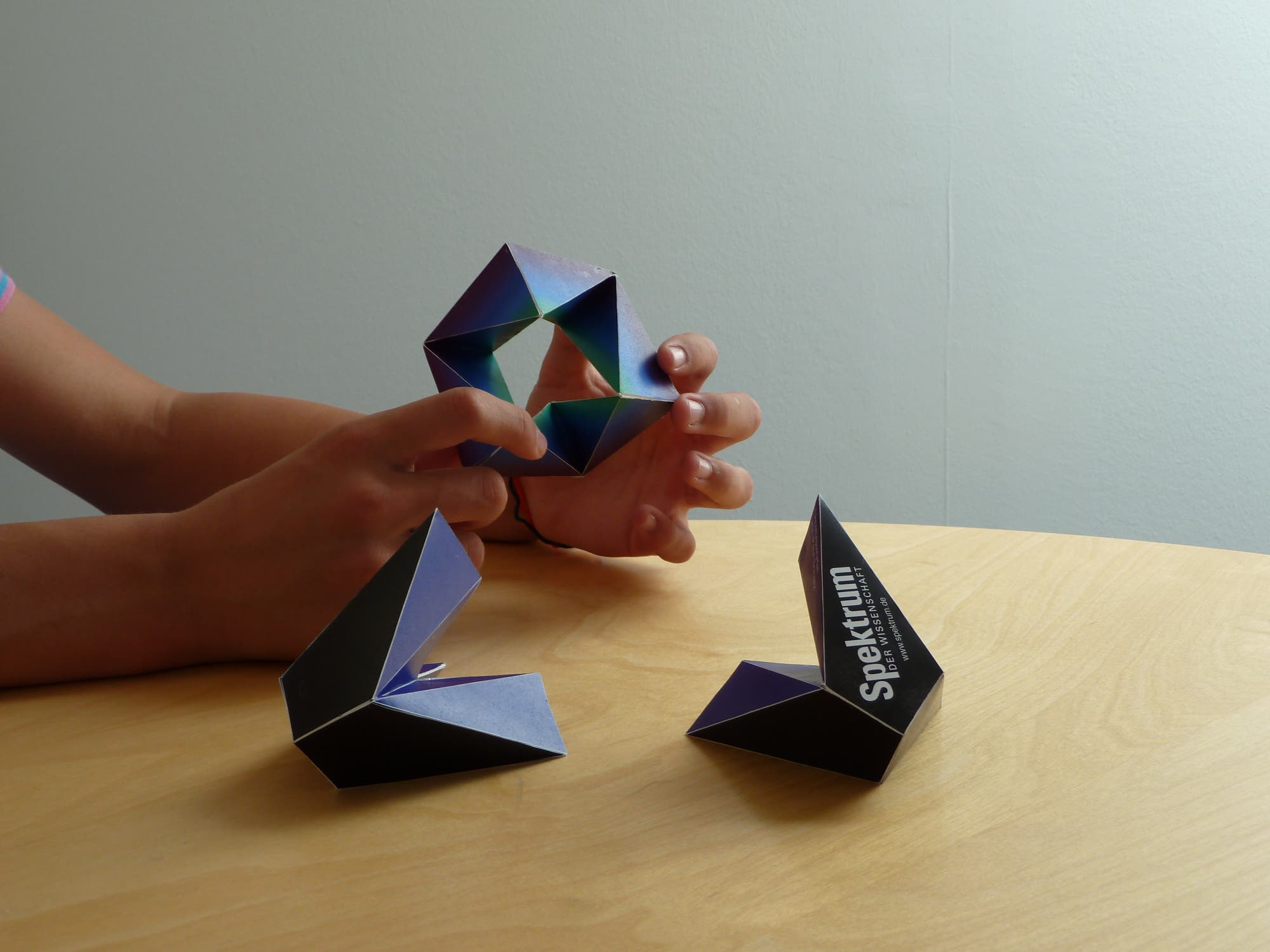 Eine Person formt mit den Händen ein geometrisches Origami-Objekt aus blauen und violetten Papierstücken. Auf dem Tisch liegen zwei weitere ähnliche Origami-Formen. Auf einer der Formen ist der Text "Spektrum" zu sehen. Die Szene spielt sich auf einem hellen Holztisch vor einer neutralen Wand ab.