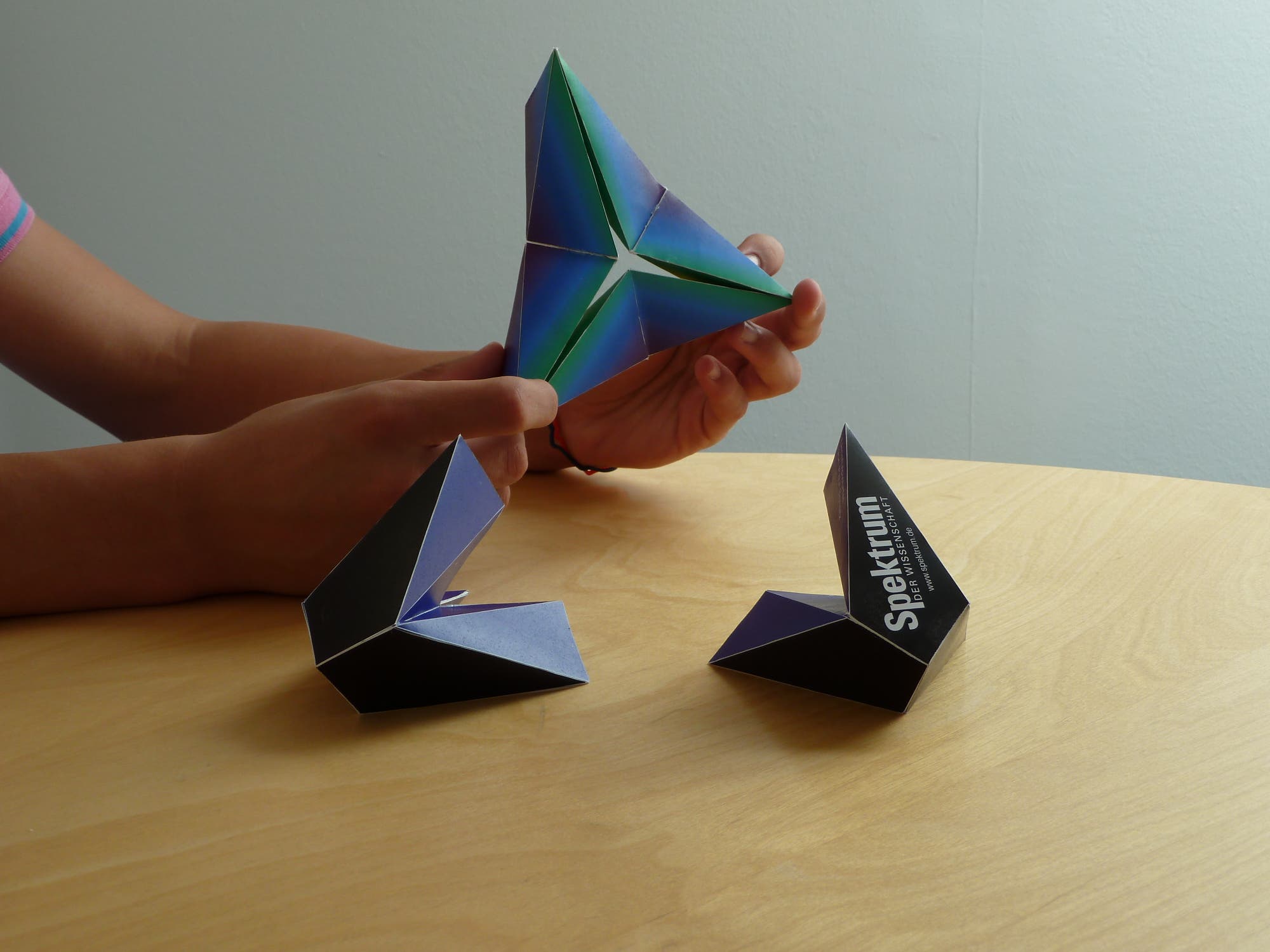 Eine Person hält ein farbenfrohes, dreieckiges Origami-Modell in den Händen. Auf dem Tisch liegen zwei weitere ähnliche Origami-Modelle. Eines der Modelle zeigt den Text "Spektrum". Die Szene spielt sich auf einem hellen Holztisch vor einer neutralen Wand ab.