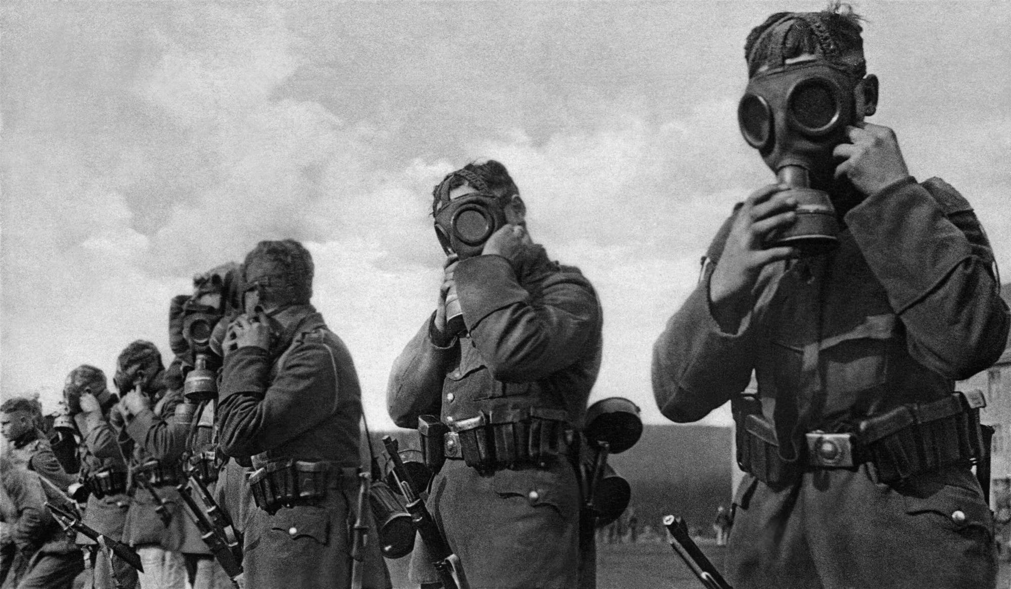 Eine Schwarz-Weiß-Aufnahme von einer Reihe Soldaten, die gerade alte Modelle von Gasmasken über ihr Gesicht ziehen und dort fixieren