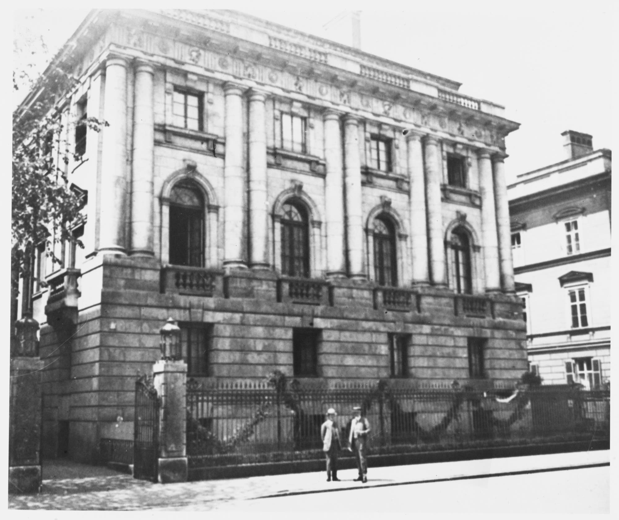 Ein historisches Gebäude mit einer Fassade aus Stein und mehreren Säulen. Vor dem Gebäude stehen zwei Personen auf dem Gehweg. Ein schmiedeeisernes Tor umgibt das Grundstück. Das Gebäude hat große Fenster und architektonische Details im klassischen Stil. Im Hintergrund rechts ist ein weiteres Gebäude auf der Schwarz-Weiß-Aufnahme sichtbar.