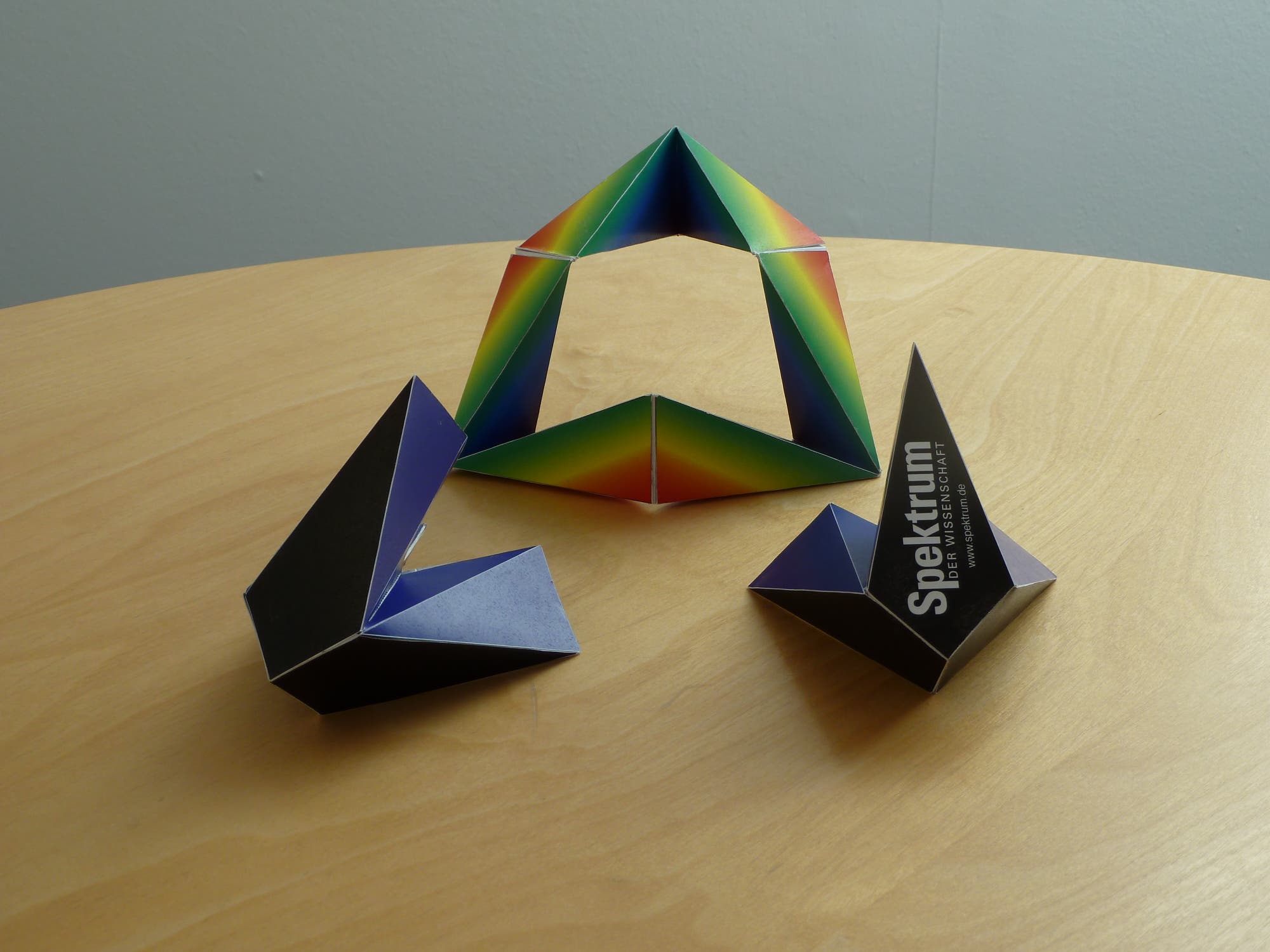 Drei bunte, geometrische Papiermodelle auf einem Holztisch. Das Modell in der Mitte hat eine Regenbogenfärbung und eine pyramidenartige Form. Die beiden anderen Modelle sind dunkler, eines davon zeigt den Text "Spektrum der Wissenschaft". Die Modelle sind kunstvoll gefaltet und präsentieren verschiedene Formen und Farben.