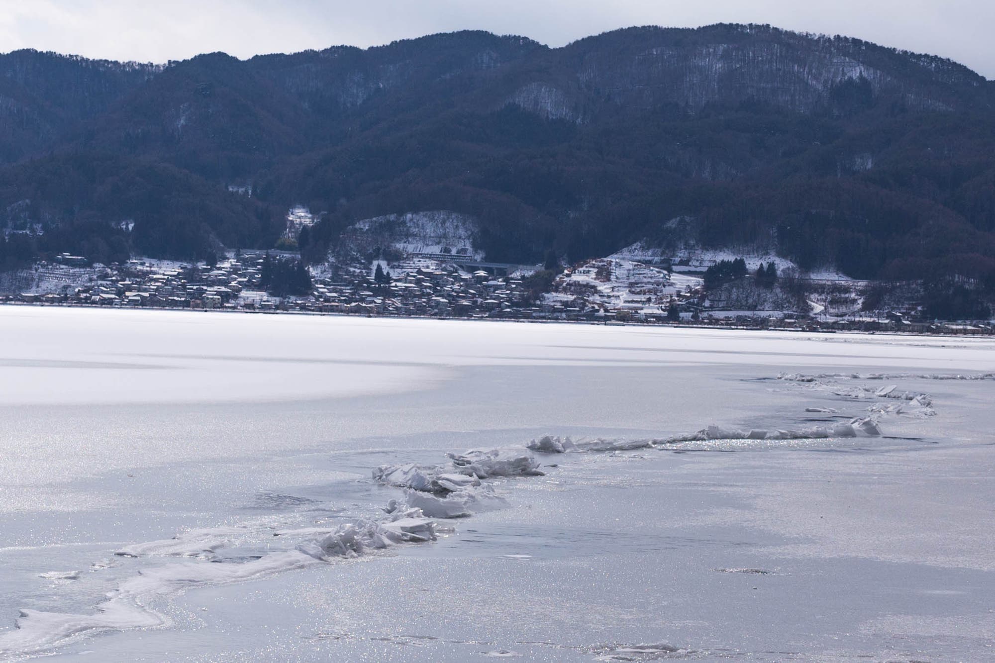 Verschneite Berglandschaft mit einem gefrorenen See im Vordergrund. Am Ufer des Sees liegt ein kleines Dorf mit schneebedeckten Dächern. Im Hintergrund erheben sich bewaldete Berge, ebenfalls mit Schnee bedeckt. Die Oberfläche des Sees zeigt Risse im Eis, die sich diagonal durch das Bild ziehen. Der Himmel ist bewölkt und die gesamte Szenerie wirkt ruhig und winterlich.