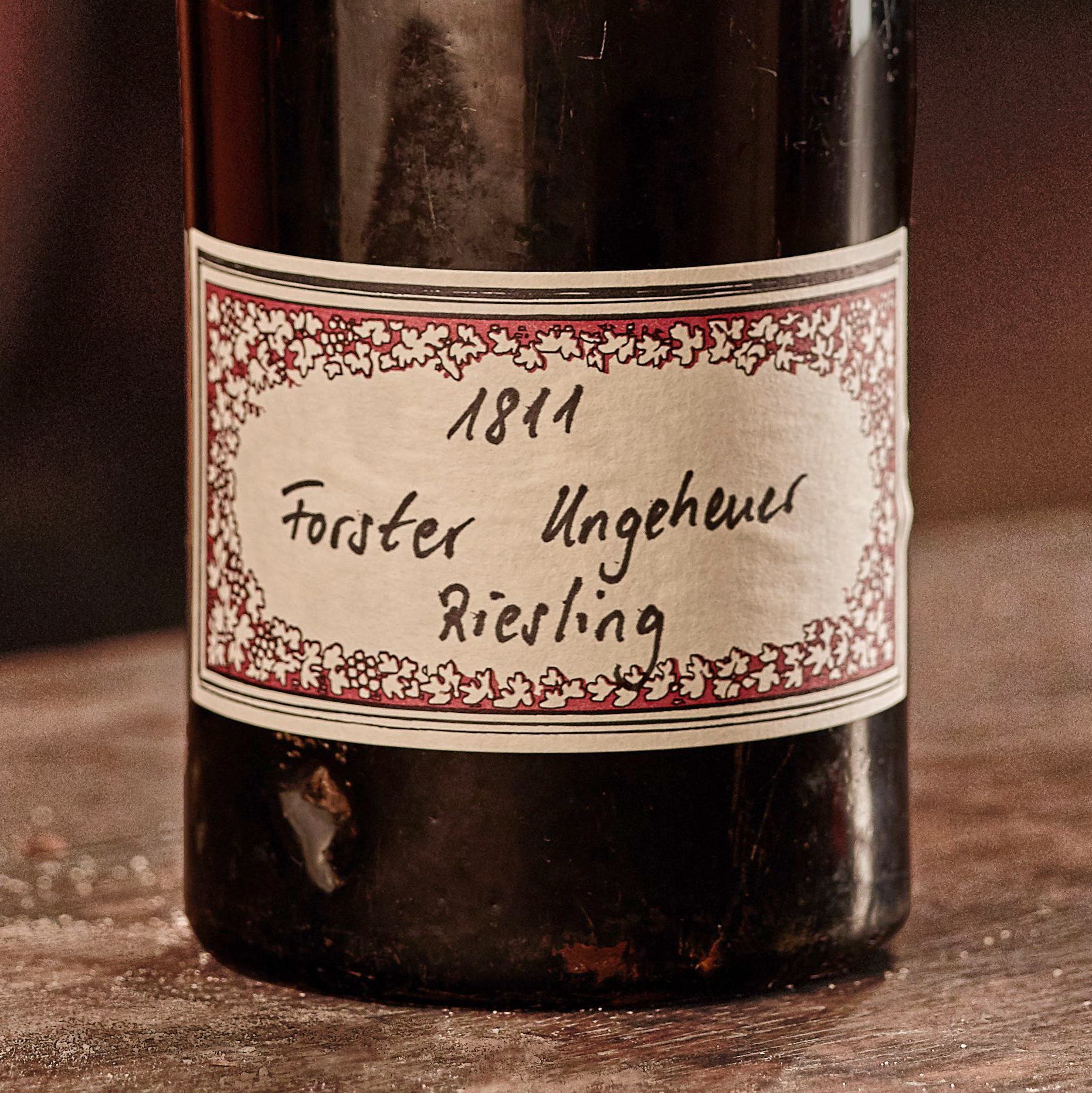 Eine Weinflasche mit einem Etikett, das handschriftlich die Aufschrift »1811 Forster Ungeheuer Riesling« trägt. Das Etikett ist von einem dekorativen roten Rand mit weißen Blättern umgeben. Die Flasche steht auf einer hölzernen Oberfläche.