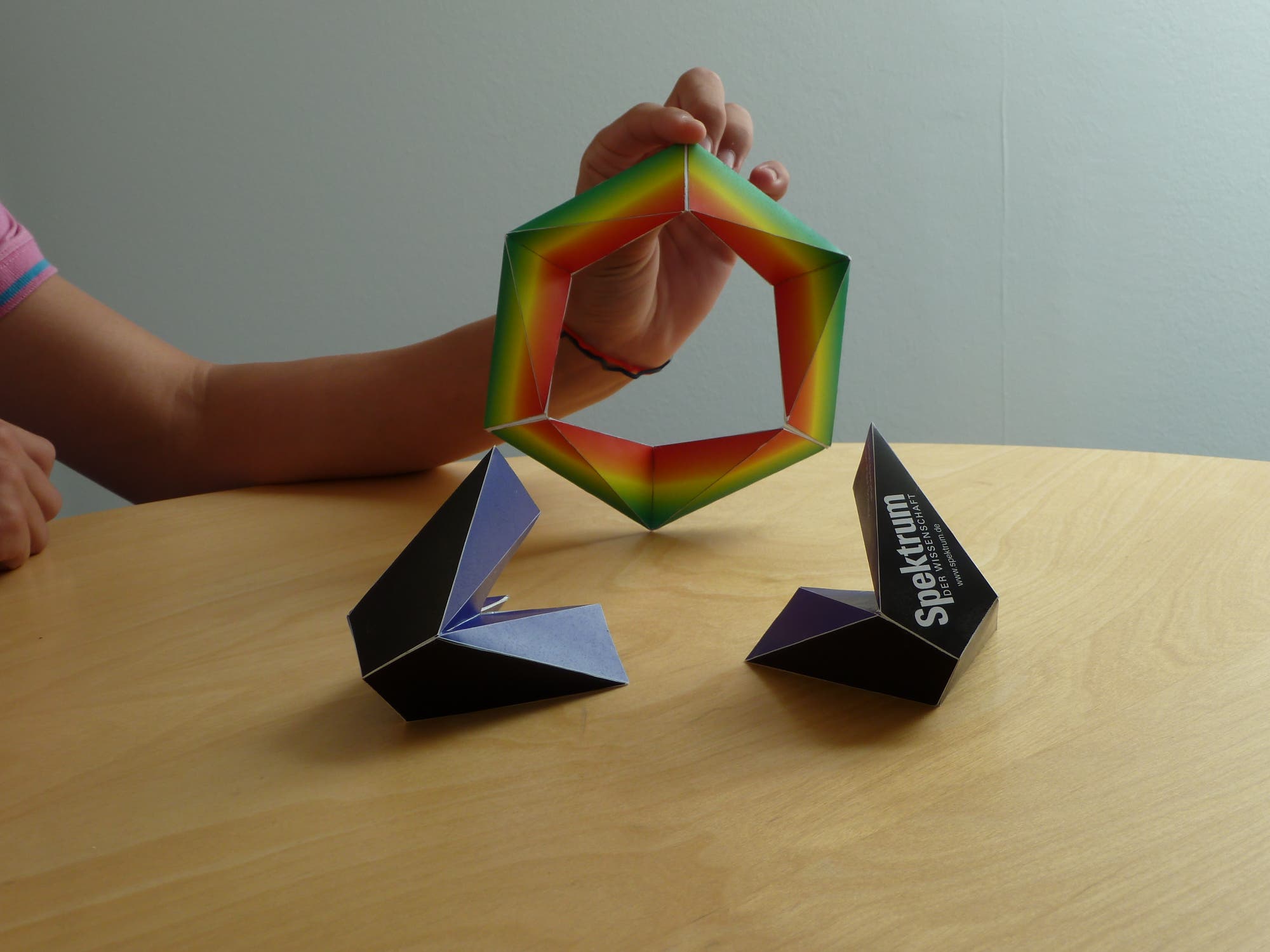 Eine Person hält ein farbenfrohes, sechseckiges Origami-Modell über einem Holztisch. Auf dem Tisch stehen zwei weitere Origami-Modelle in dunklen Farben, eines davon mit dem Text "Spektrum" sichtbar. Die Szene zeigt die kreative Kunst des Papierfaltens.