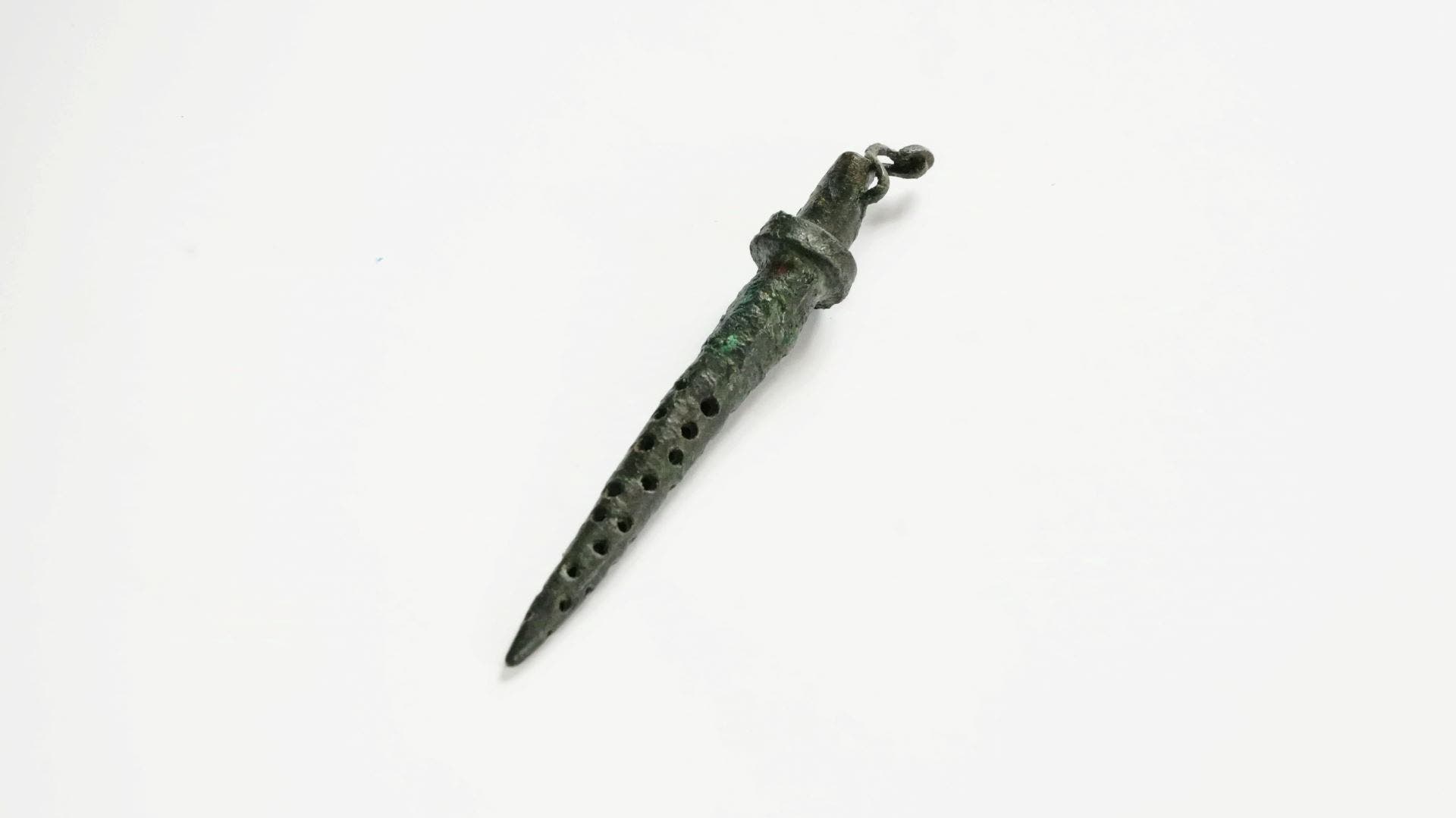 Ein antikes bronzenes Blasinstrument, das wie eine Flöte geformt ist, liegt auf einem weißen Hintergrund. Das Instrument hat mehrere Löcher entlang des Korpus und einen verzierten Griff. Es scheint aus der Bronzezeit zu stammen und könnte in historischen oder archäologischen Kontexten von Interesse sein.