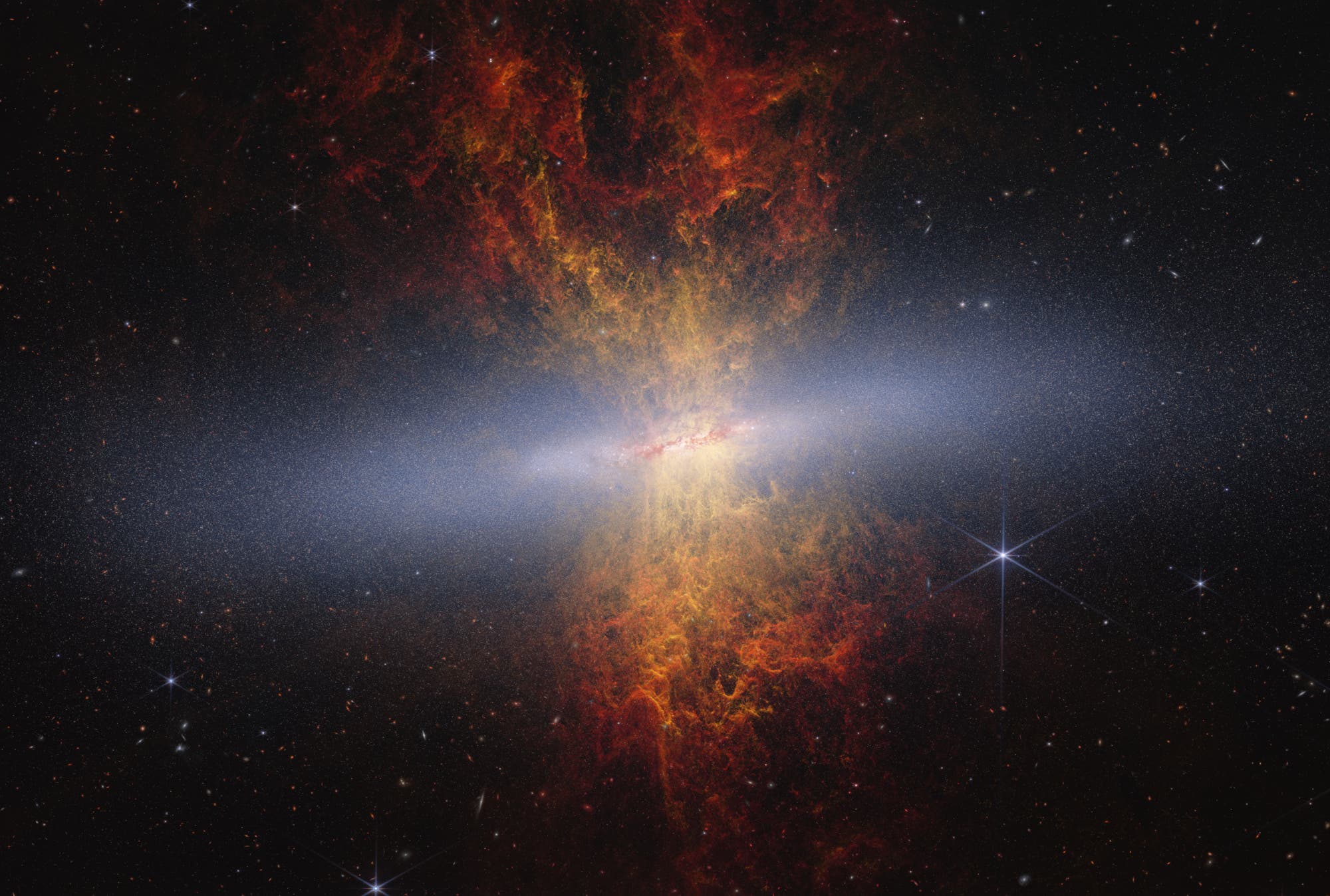 Eine beeindruckende astronomische Aufnahme zeigt eine Galaxie mit einem hellen, zentralen Kern, umgeben von einem diffusen, leuchtenden Nebel in Rot- und Gelbt&ouml;nen. Die Galaxie erstreckt sich horizontal &uuml;ber das Bild, w&auml;hrend der Nebel vertikal aus dem Zentrum herausragt. Im Hintergrund sind zahlreiche Sterne und Galaxien zu sehen, die den tiefen, dunklen Weltraum darstellen. Die Aufnahme vermittelt ein Gef&uuml;hl von Weite und Komplexit&auml;t des Universums.