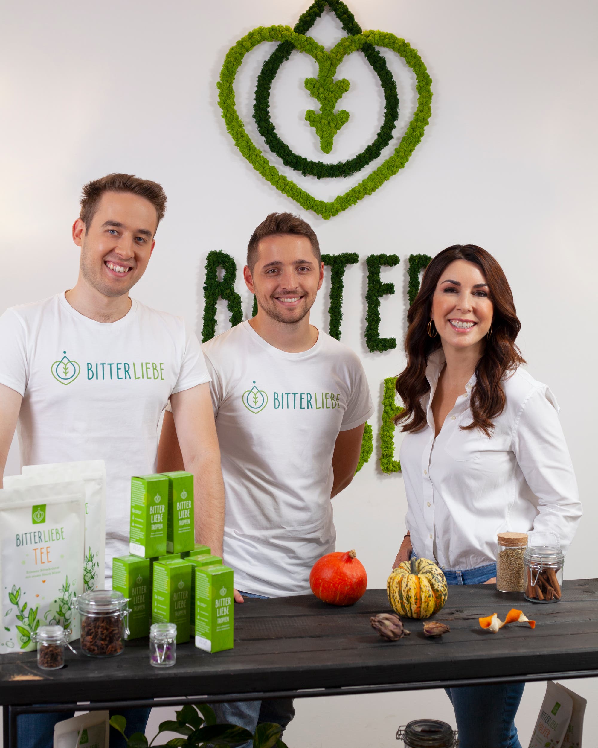 Jan Stratmann, Andre Sierek und Judith Williams vor einer Wand mit dem Logo von BitterLiebe. Auf einem Tisch vor ihnen sind verschiedene Produkte von BitterLiebe zu sehen, darunter BitterLiebe Tee und BitterLiebe Tropfen sowie Gewürzgläser und Kürbisse.