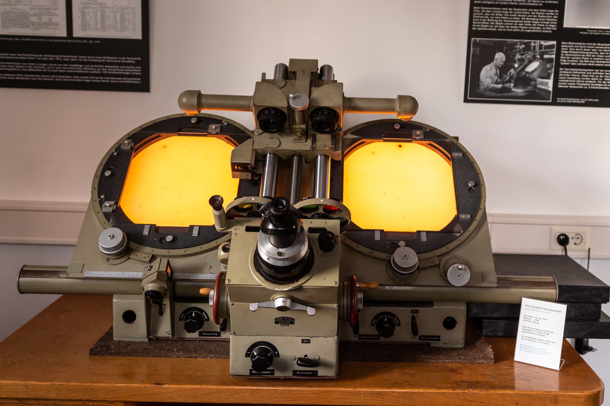 Ein altes optisches Gerät, ein Plattenkomparator, mit zwei beleuchteten Bildschirmen steht auf einem Holztisch. Das Gerät hat mehrere Drehknöpfe und Linsen in der Mitte. Neben dem Gerät steht rechts eine kleine Informationstafel.
