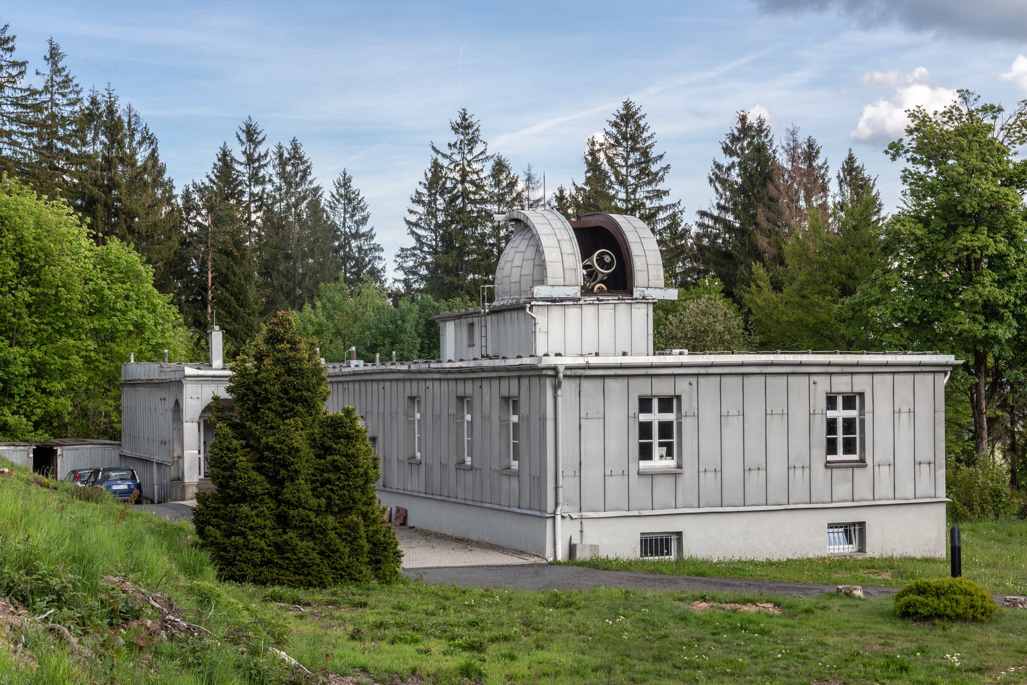 Das Alte Hauptgebäude ist ein Observatoriumsgebäude mit einer geöffneten Kuppel, die ein Teleskop zeigt. Das Gebäude ist von Bäumen umgeben und steht auf einer grünen Wiese. Der Himmel ist leicht bewölkt. Im Hintergrund sind hohe Nadelbäume zu sehen.