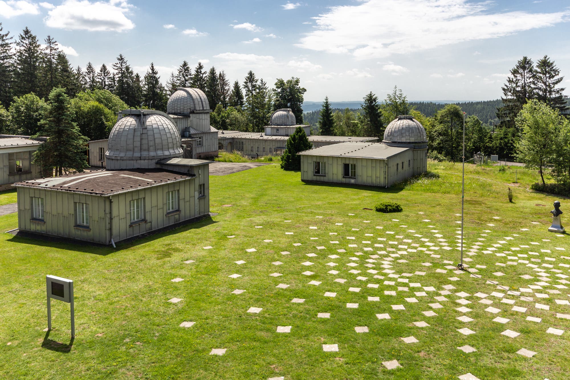 Das Foto zeigt einen Teil der Gebäude an der Sternwarte Sonneberg, einige von ihnen mit Kuppeldächern. Sie stehen auf einer grünen Wiese, umgeben von Bäumen. Im Vordergrund sind geometrisch angeordnete Steinplatten auf dem Rasen zu sehen: der SkyPole. Der Himmel ist leicht bewölkt, und die Szene vermittelt eine ruhige, wissenschaftliche Atmosphäre. Ganz rechts steht die Büste eines Mannes.