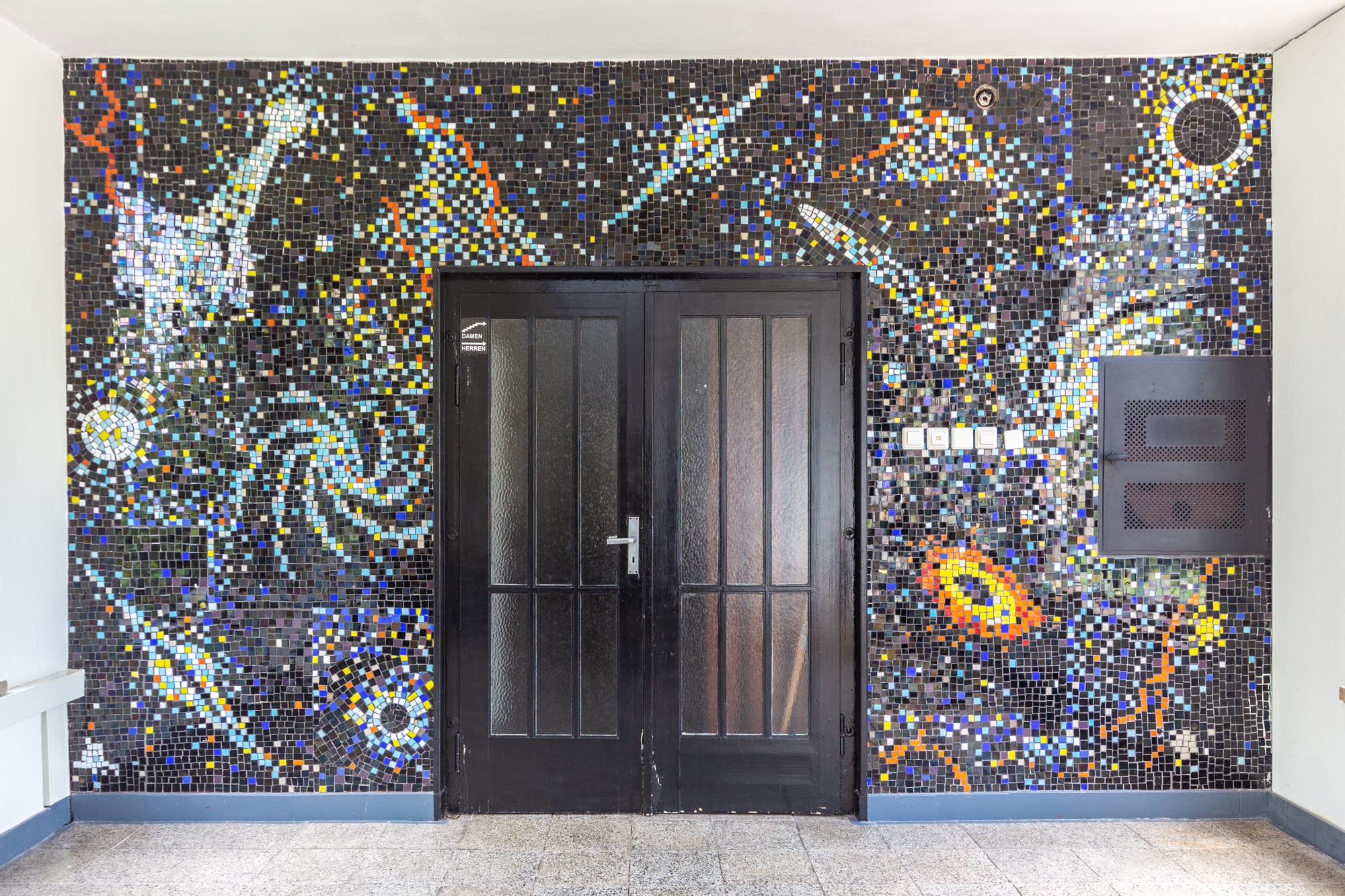Eine Wand mit einem farbenfrohen Mosaik, das kosmische Motive darstellt. Das Mosaik zeigt Sterne, Galaxien und Planeten in leuchtenden Farben auf einem dunklen Hintergrund. In der Mitte befindet sich eine Doppeltür, die von dem Mosaik umrahmt wird. Rechts neben der Tür sind Schalter und ein Kasten sichtbar. Das Kunstwerk vermittelt ein Gefühl von Weite und Unendlichkeit des Universums.